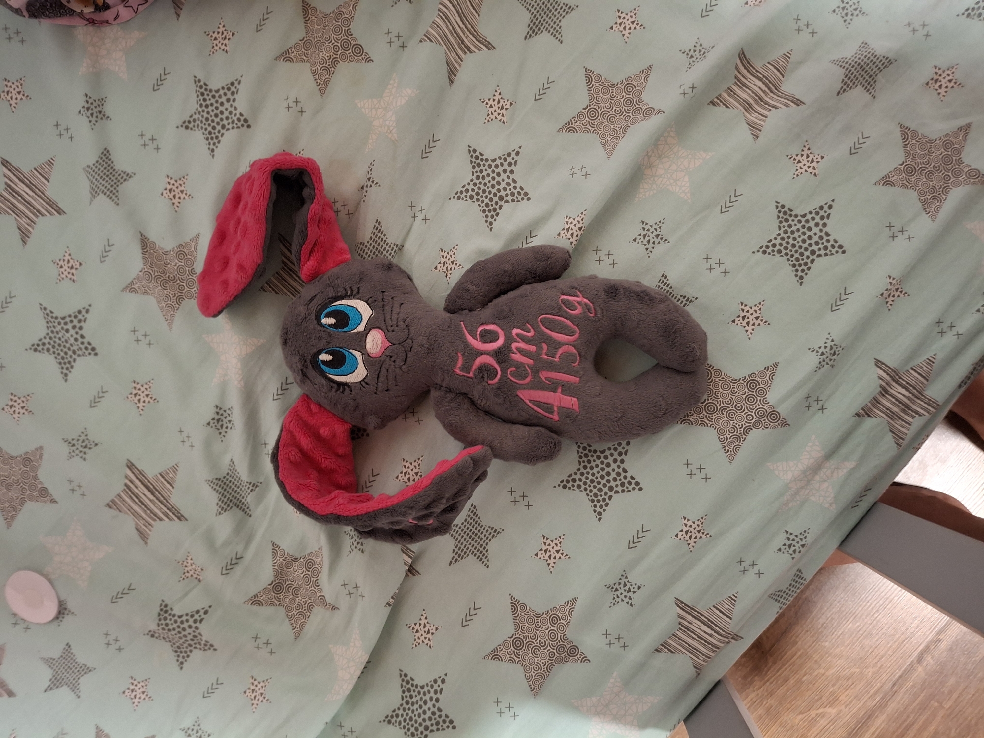 This media file shows a close-up, overhead shot of a plush stuffed bunny toy lying on a mint green fabric covered in a pattern of stars and small crosses. The bunny is gray with large, blue cartoonish eyes, pink inner ears, and pink accents on its feet. Embroidered on its stomach in pink thread are the numbers "56" followed by "cm" and "4150g". The fabric appears to be a baby blanket or bedding, given the gentle pattern and the presence of the stuffed toy. The lighting suggests an indoor setting, possibly a nursery or a child's room, and the absence of natural light cues implies it could be any time of day. The overall tone is peaceful and domestic, highlighting a baby-related item. No people are visible.