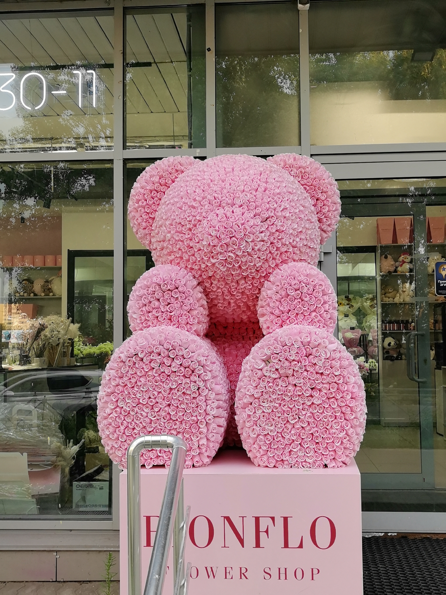 The image shows a large, pink teddy bear made of roses displayed outside a shop. The shop's name, "BONFLO FLOWER SHOP," is visible on a pink pedestal beneath the teddy bear. The shop appears to be a flower shop, as indicated by the name and the presence of flowers visible through the glass window behind the teddy bear. The shop has a modern storefront with large glass windows and a neon sign displaying "30-11" in white. The exterior of the shop suggests an urban environment. The teddy bear is the primary subject, a large, intricately constructed floral arrangement. The scene is well-lit, possibly during daylight hours, with reflections visible on the shop windows.