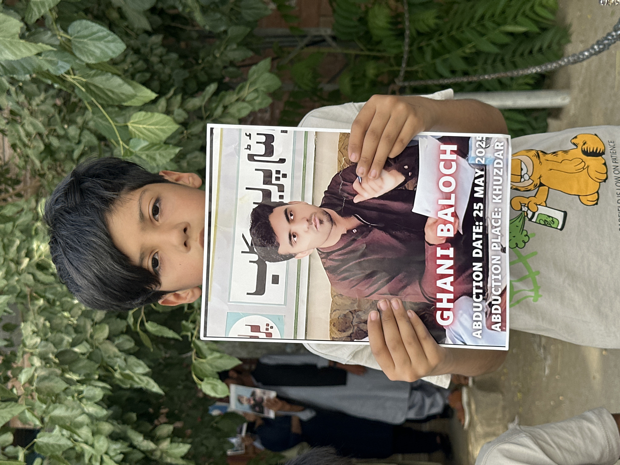 A young boy holds a poster with a picture of Ghani Baloch, stating the abduction date of May 25, 2025, and the abduction place as Khuzdar. The scene appears to be an outdoor demonstration or gathering, as people holding similar posters can be seen in the background. The location is characterized by green foliage.