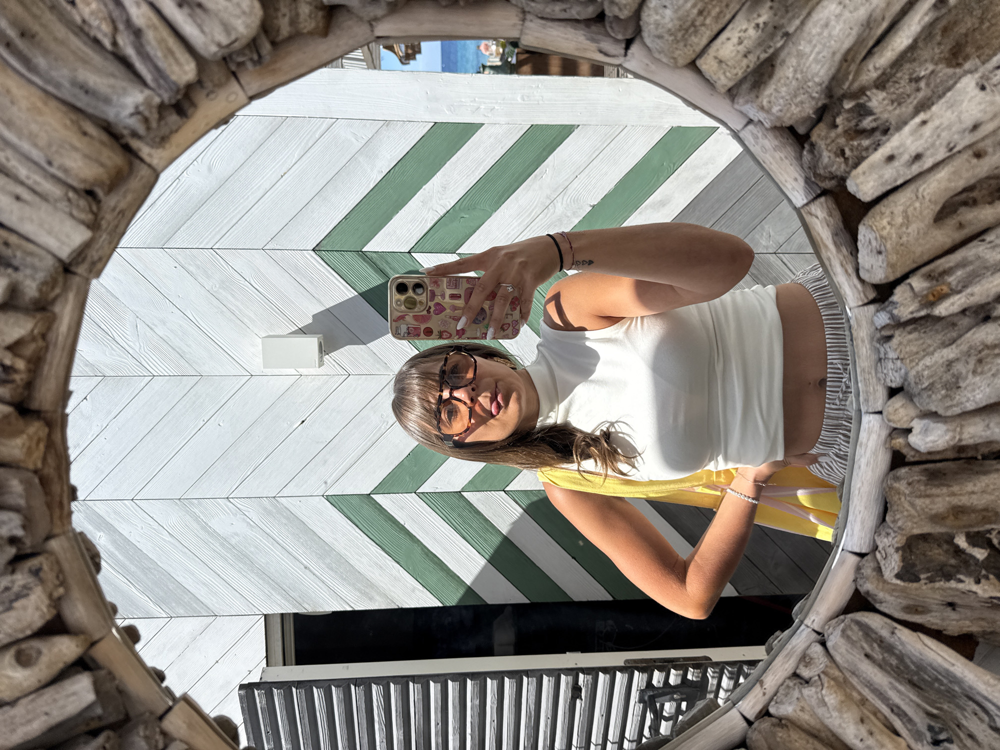 Captured through a circular, driftwood-framed mirror, the scene portrays a woman taking a selfie in what appears to be a stylish establishment. The location is specified as Ramatuelle, France. The woman, wearing glasses and a white top with a yellow accessory, holds her phone up to capture her reflection. The background consists of a chevron-patterned wall, alternating between white and green. Below the woman, a black window and part of a corrugated metal railing are visible. The lighting is bright and sunny.