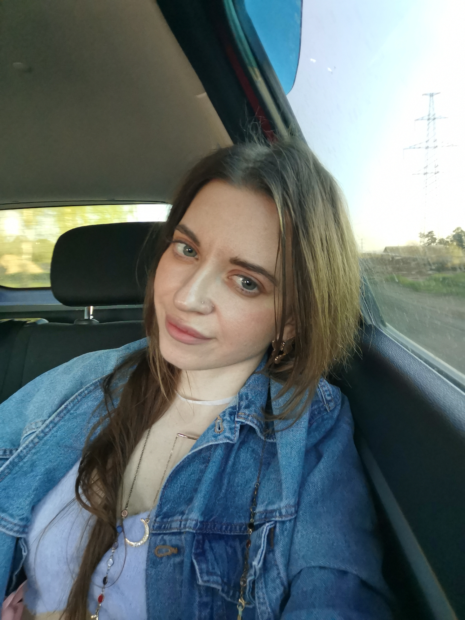 A woman is sitting in the back seat of a car. She has light brown hair, light skin, and is wearing a blue denim jacket over a light purple top. She has several necklaces on. Through the car window, electrical pylons and some greenery can be seen. The location is Chaikovsky, Russia.