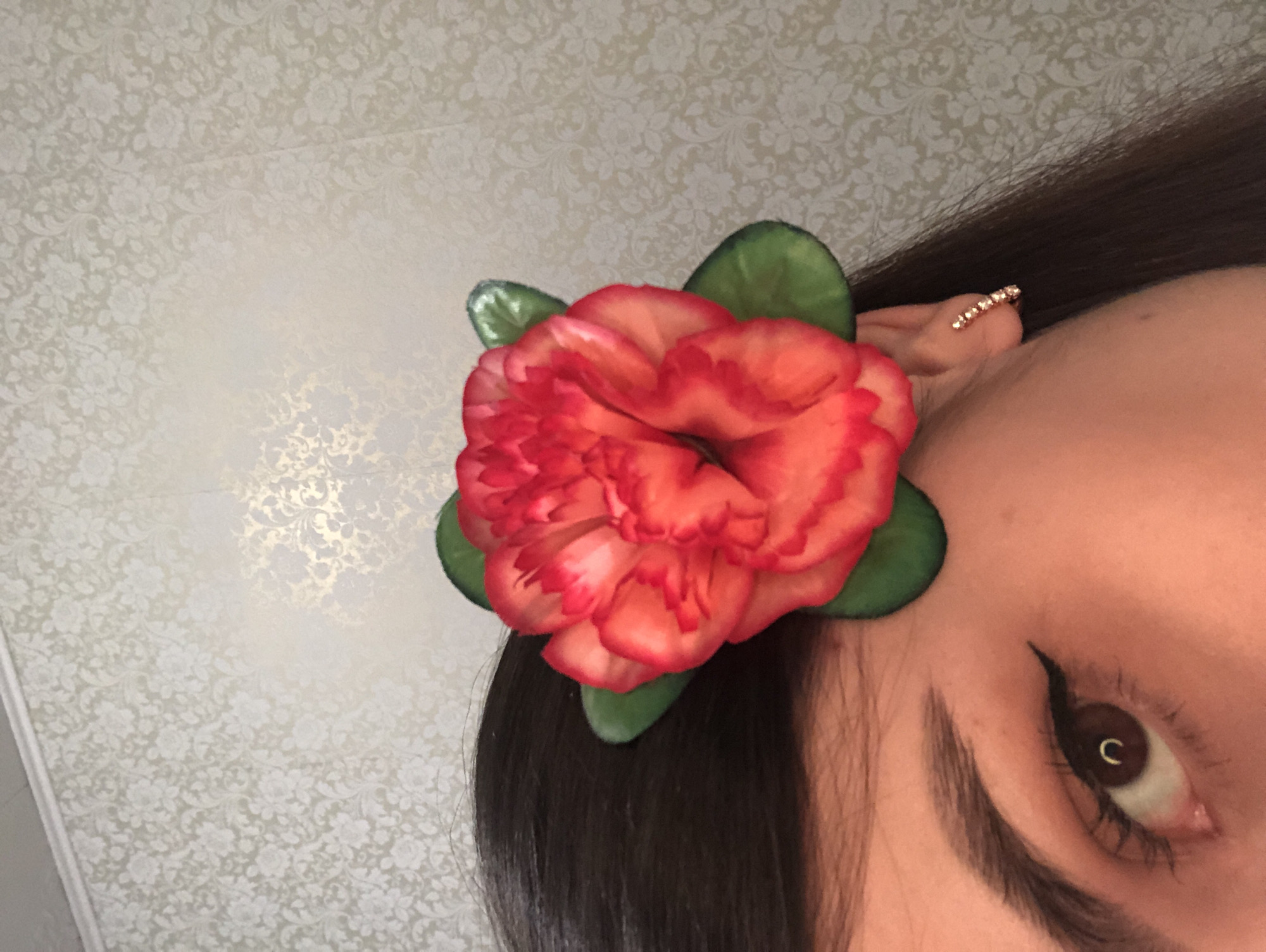 A close-up, angled shot features a person with dark hair. An orange flower with green leaves is positioned near their ear. They have an earring with stones. The person's eye is visible, showing a lined upper eyelid. The background is a light-colored wallpaper with a floral pattern. The scene is not identifiable in the city of Noviy, Russia.