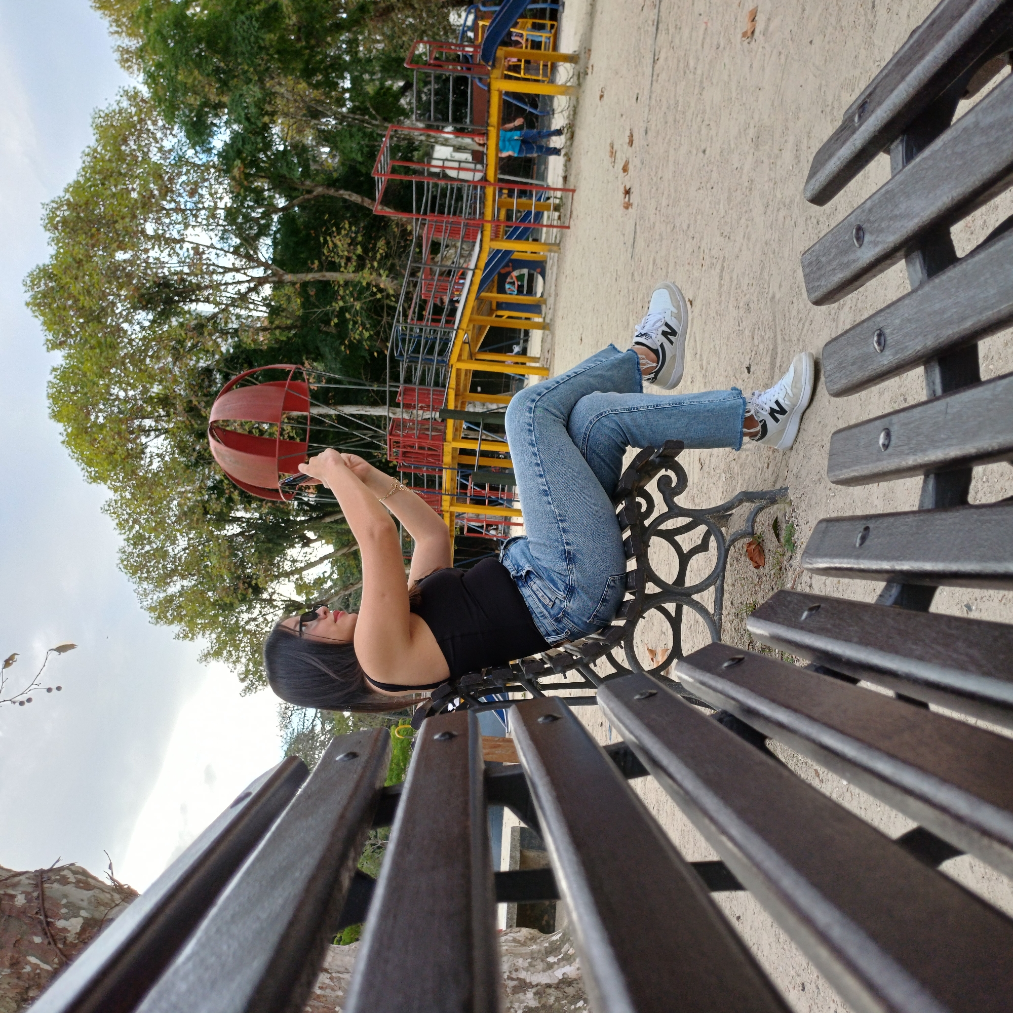 The media file, presented on its side, captures a person relaxing horizontally on a dark-colored park bench. The person, with dark hair and wearing sunglasses, a black tank top, light blue ripped jeans, and white sneakers with a visible "N" logo, has their legs crossed and hands raised as if holding a smartphone or camera. A thin gold bracelet is visible on their right wrist.
In the background, to the right of the person, a colorful playground structure with yellow, red, and blue components, including slides and climbing frames, is discernible. Near the person's hands, a distinctive red spherical, cage-like structure is present. Above the person and playground, lush green trees fill much of the scene, while a bright, possibly overcast sky is visible in the background, to the left. The ground, appearing sandy or dusty with scattered dry leaves, stretches from beneath the bench to the right side of the image. The setting suggests an outdoor park or playground in Curitiba, Brazil, during daytime. The person appears relaxed, engaging in a leisure activity.