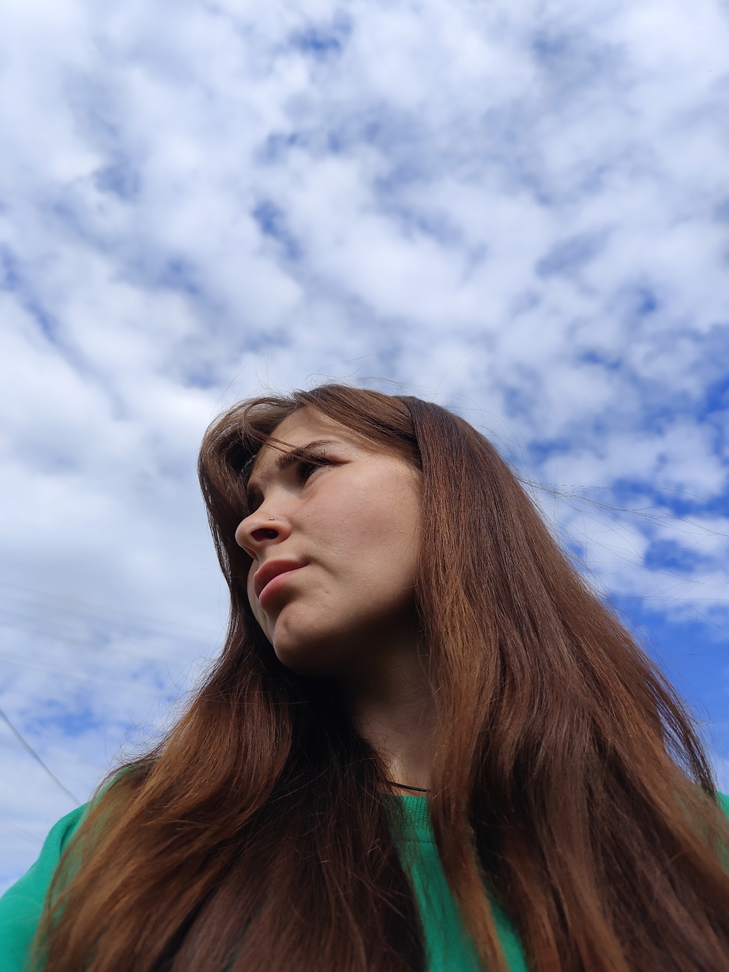 A young woman is depicted outdoors in Фоки, Russia, viewed from a low angle, looking upwards and to the upper left of the frame. She has long, straight to slightly wavy brown hair with reddish undertones, which appears lightly wind-blown, with individual strands visible. Her left profile and a three-quarter view of her face are presented, showing fair skin, a small gold-colored stud piercing in her left nostril, and a neutral, slightly pensive expression as she gazes towards the sky. She wears a plain emerald green crew-neck top. The background consists of a clear blue sky interspersed with numerous scattered white cumulus clouds. In the far left background, three faint, thin, dark horizontal lines, consistent with utility wires, are visible against the sky.