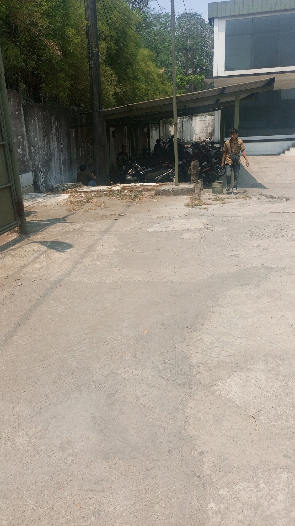 The image depicts an outdoor scene in Kadu, Indonesia, focusing on a paved area with a covered structure housing several motorcycles. In the foreground, a light-colored concrete surface with visible cracks and debris suggests an industrial or utilitarian setting. To the left, a portion of a green metal gate is visible. A tall, dark utility pole stands prominently in the mid-ground, with wires extending from it. Behind the utility pole and to the left, a weathered concrete wall borders the scene, topped with lush green bamboo foliage.
Under a covered structure, numerous motorcycles are parked in rows. Two individuals are present within this area. One person, seated and facing away from the camera, is wearing a dark shirt and appears to be looking at the motorcycles. Another person, a young male wearing a light brown t-shirt and dark pants, is walking from right to left across the paved area. He carries a dark bag slung across his body.
In the background, a modern building with large, dark windows and white trim is visible on the right. The overall lighting suggests a sunny day. The environment appears somewhat unkempt, with dried leaves and small patches of grass growing in the paved areas. The scene suggests a parking area or a facility in Kadu, Indonesia, possibly related to a business or transportation hub, given the presence of motorcycles and the industrial-style building.