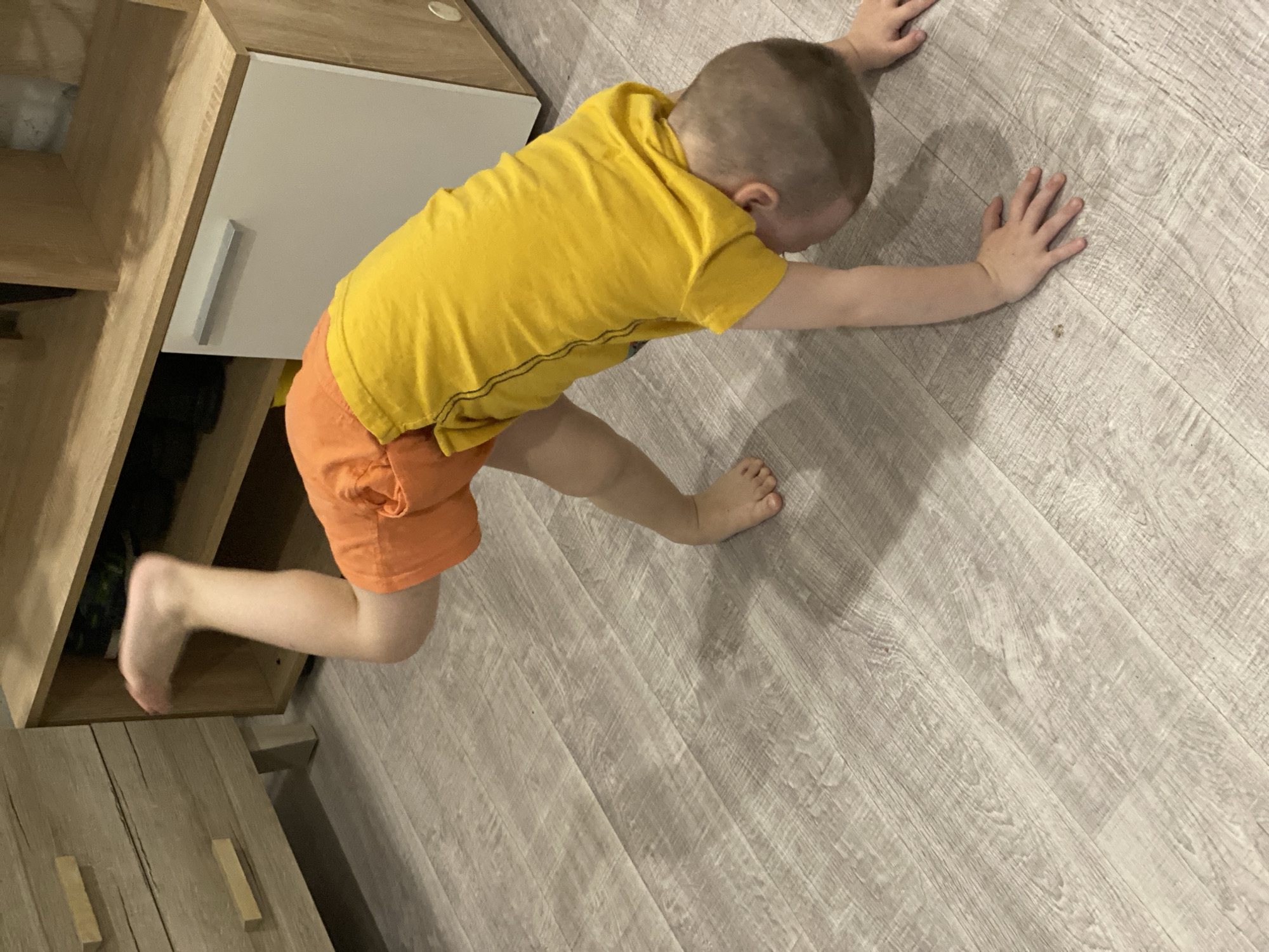 A young child, wearing a yellow shirt and orange shorts, is lying on a light grey wood-patterned floor. The child extends both arms forward and presses their hands on the floor. The child's head is turned downward towards the floor. One foot is on the floor while the other is extending into a partially visible wooden cabinet with drawers. The environment appears to be indoors. Sochi, Russia is not discernible from the image.