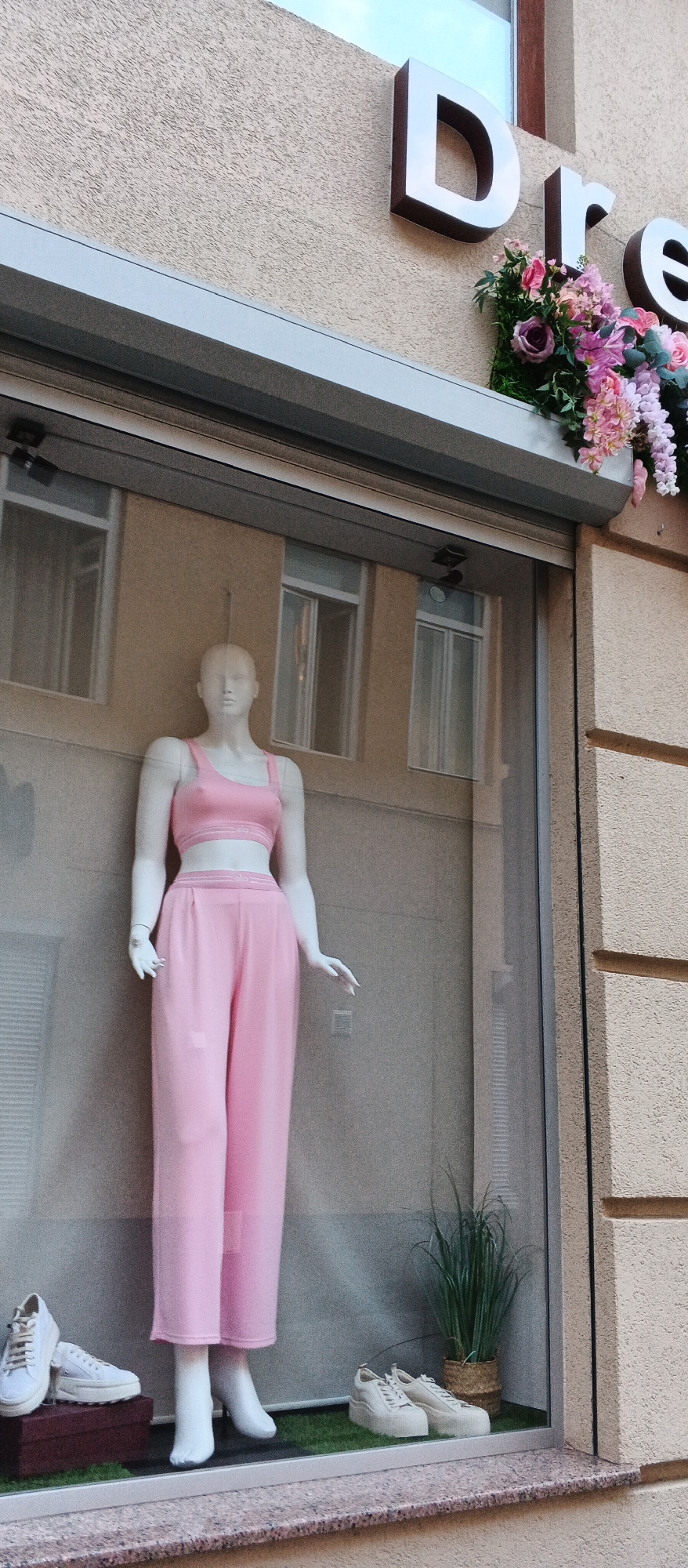 The image is a vertical, eye-level, medium shot of a shop window display in Mukachevo, Ukraine. A female mannequin, positioned centrally, wears a pink two-piece athleisure outfit consisting of a sports bra and wide-leg pants. The mannequin has white skin and no facial features. The clothing appears to have a subtle striped logo on the waistband of the pants and the band of the sports bra.
To the left of the mannequin, on a dark red box, are two white sneakers. To the right of the mannequin, on a patch of artificial green grass, are two white platform sneakers, and a potted artificial green plant with long, thin leaves in a woven basket.
Behind the mannequin, the shop window reflects the interior of the store and the exterior facade of the building opposite. The shop's signage, visible at the top of the image, features the letters "D", "r", and "e" in large, three-dimensional metallic lettering. A floral arrangement of pink and purple flowers is displayed above and to the right of the signage. The building facade outside the window is a textured beige color. The foreground consists of a granite-like windowsill.