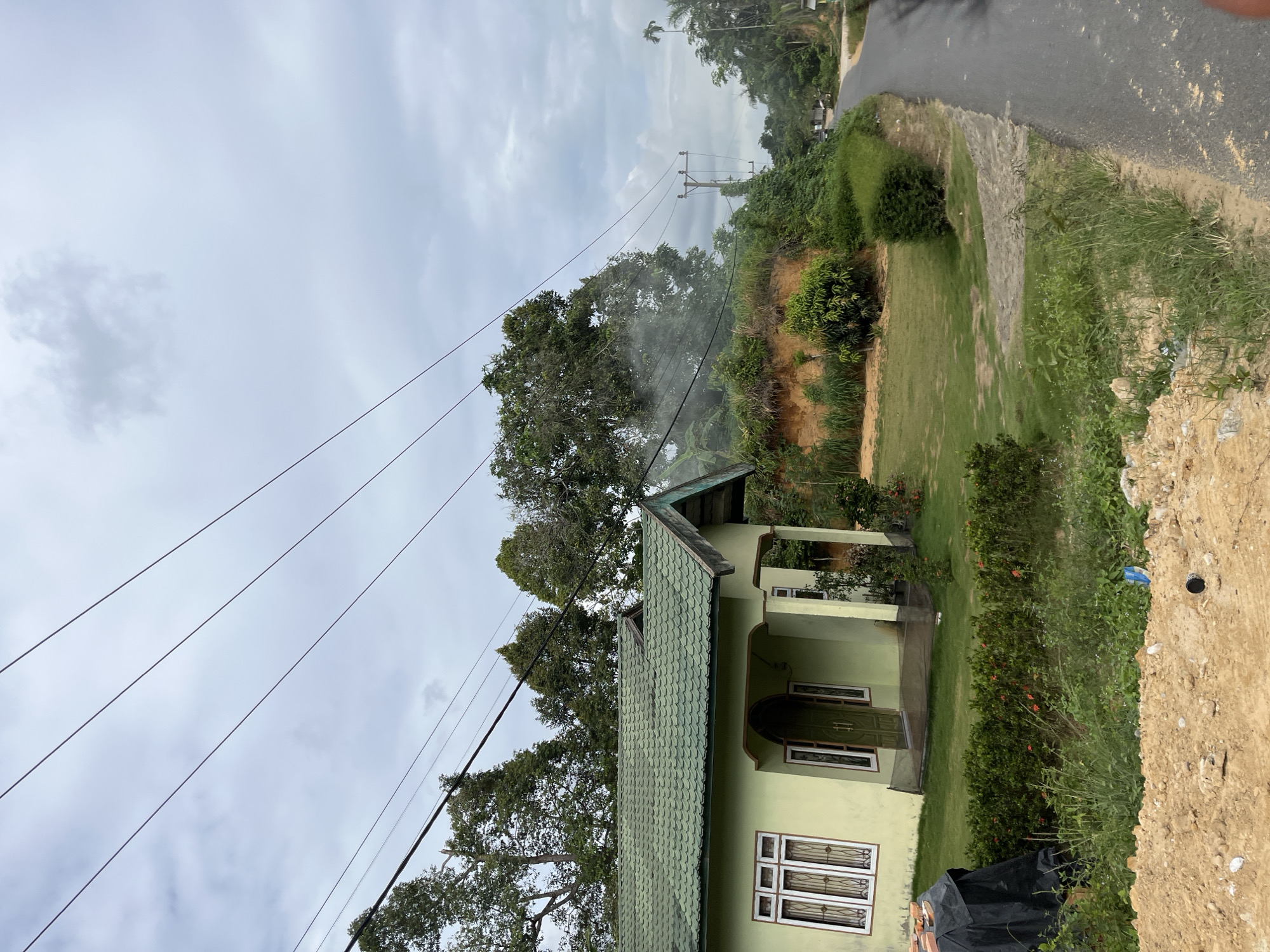A house with a light green exterior and a green tiled roof is visible in a suburban or rural setting. The house has several windows with white frames and decorative grills, and a covered porch area with a green door. Power lines run diagonally across the scene, with a telephone pole visible in the distance. The surrounding environment is lush and green, with many trees and dense vegetation. A paved road runs alongside the property, with a sandy embankment to the right. The sky is overcast with grey clouds, suggesting a cloudy day. There is a patch of mist or smoke emanating from the trees near the telephone pole. A black tarp is visible in the foreground on the right side of the image. The location is identified as Sukajadi, Indonesia.
