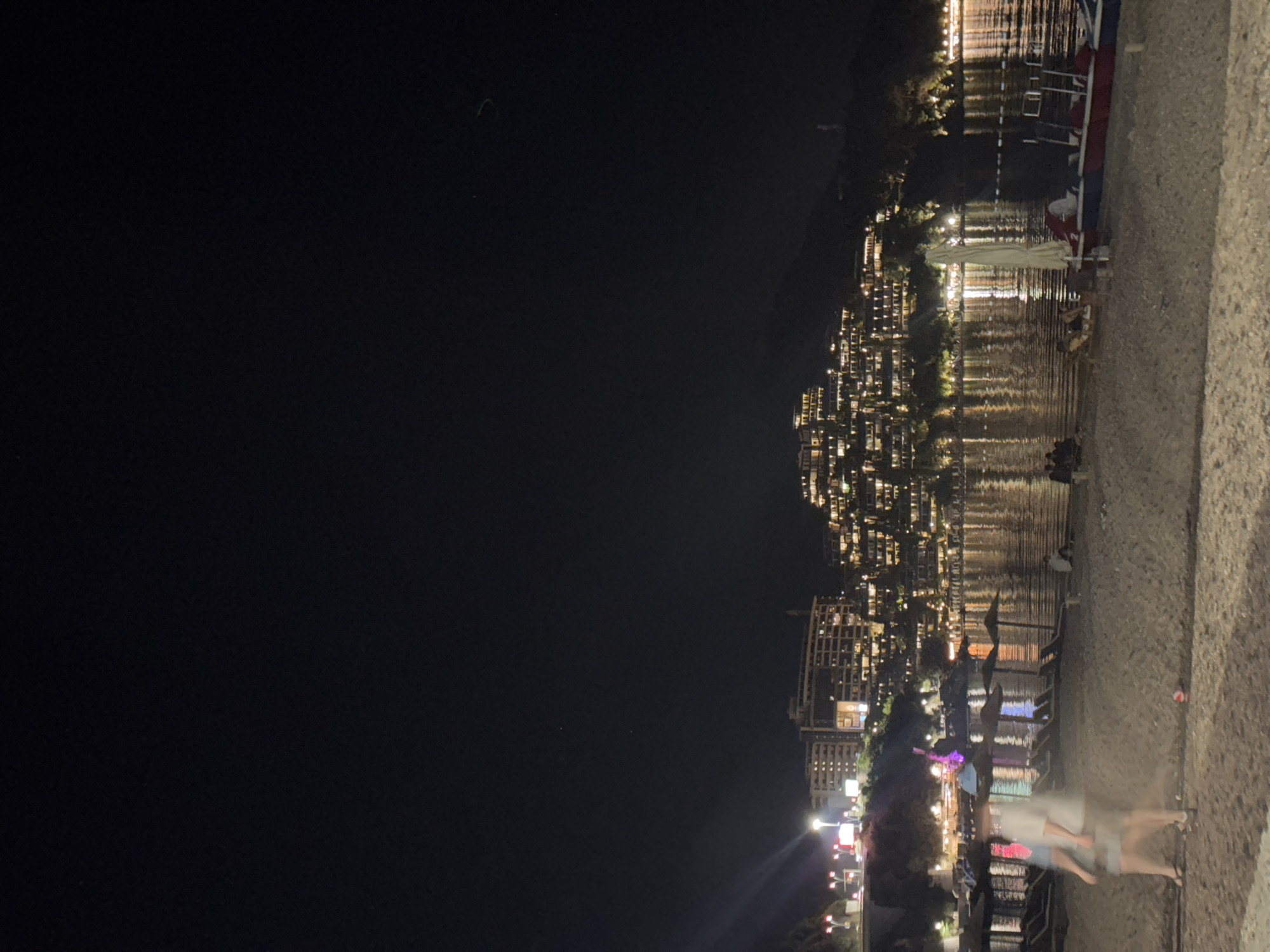 The image depicts a nighttime scene on a beach in Budva, Montenegro. In the foreground, a sandy beach is visible with several lounge chairs and umbrellas. Two blurred figures, appearing to be walking, are present on the sand. Further back, the water of the bay reflects the lights from the city. Across the bay, a hillside is covered with numerous buildings illuminated by bright lights, creating a cascading effect of light against the dark hillside. The city lights extend along the coastline. A portion of a boat or pedal boat is visible in the lower right corner of the frame, partially submerged in the water. The overall atmosphere is one of a vibrant, illuminated coastal city at night.