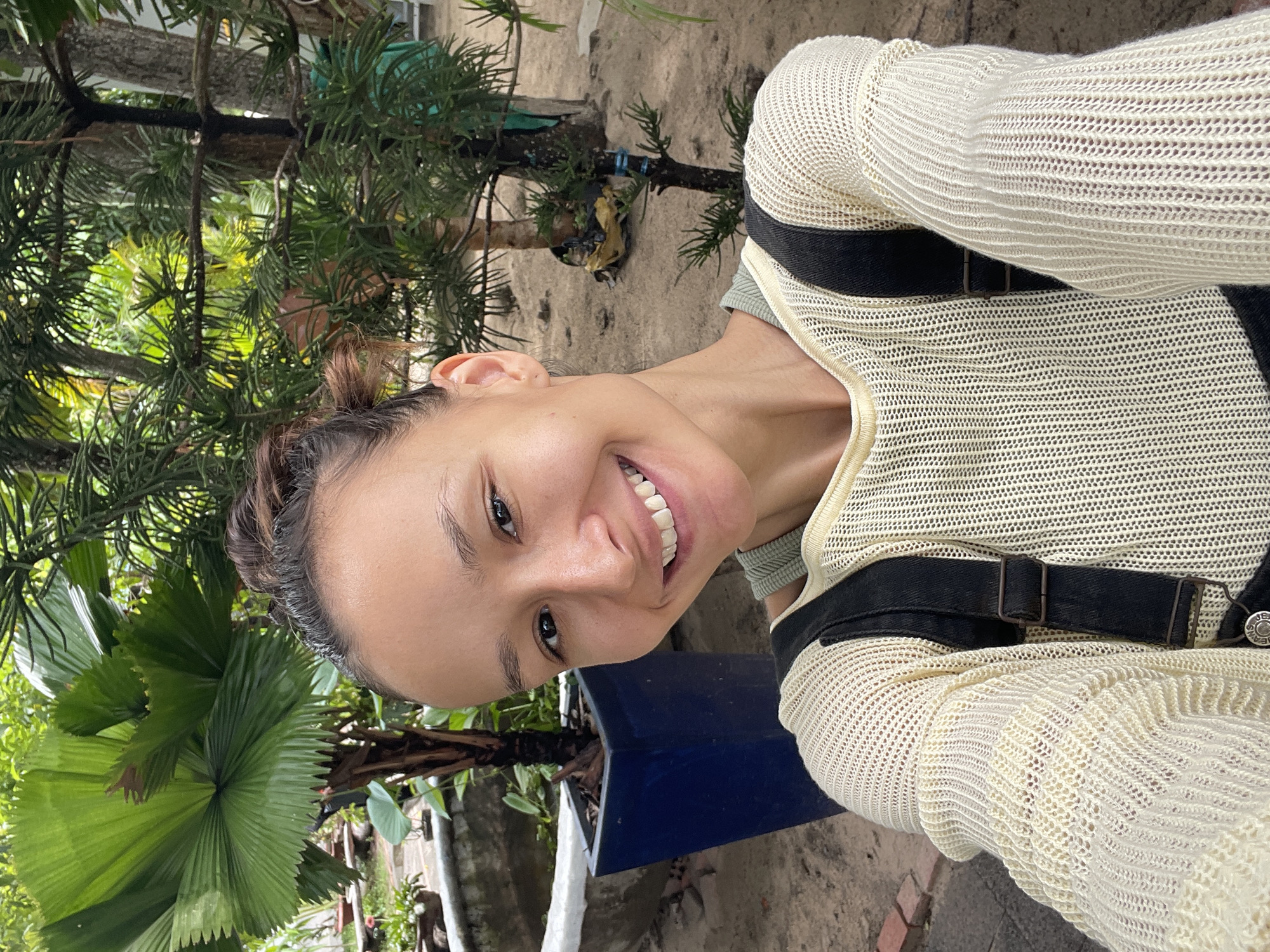 A woman with her hair up smiles in a selfie taken outdoors in what appears to be a garden or yard. She wears a beige, open-knit sweater over a light green top, and a black belt with a rectangular buckle around her waist. Green plants, including a large fan palm and coniferous-looking trees, are visible behind her. A blue rectangular container sits to her right. The location is inferred to be Phu Thuy Ward, Vietnam, based on the request.
