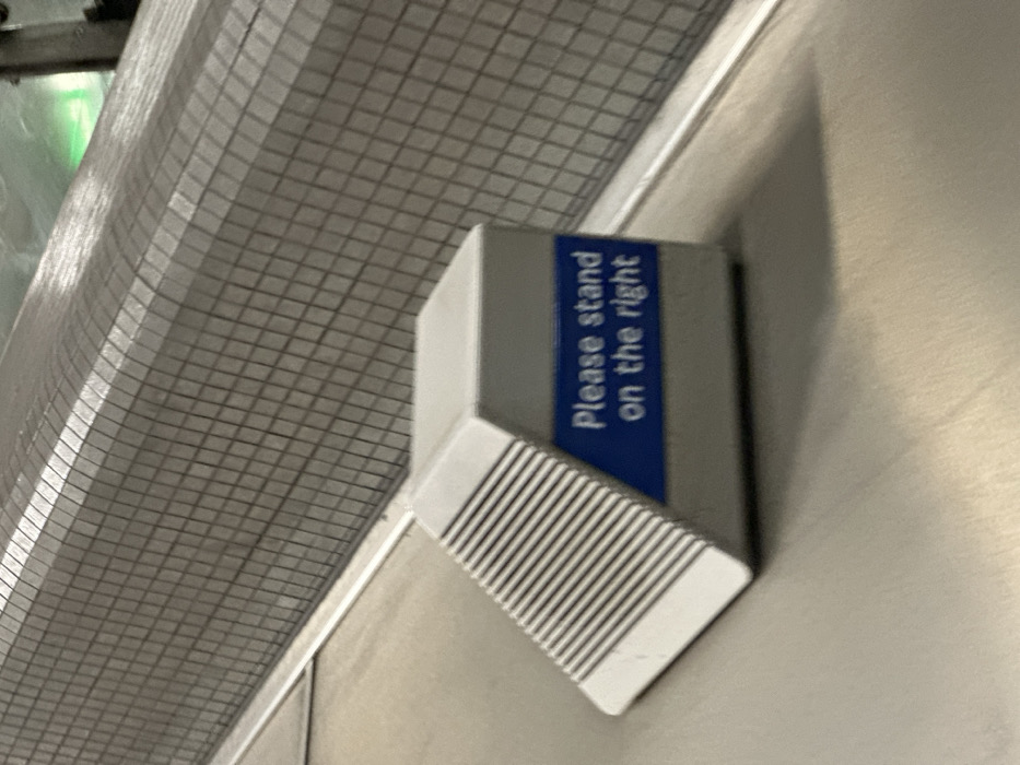 The image displays an instructional sign situated on the metallic surface of an escalator within a London Underground station in the United Kingdom. The sign is a light grey, multi-faceted object with a prominent blue horizontal band containing white sans-serif text that reads, "Please stand on the right." The sign casts a subtle shadow to its rear on the escalator surface. In the background, a curved wall, tiled with small, light grey squares, rises alongside the escalator. A faint green light source is visible in the upper right portion of the background. No individuals are present in the scene.