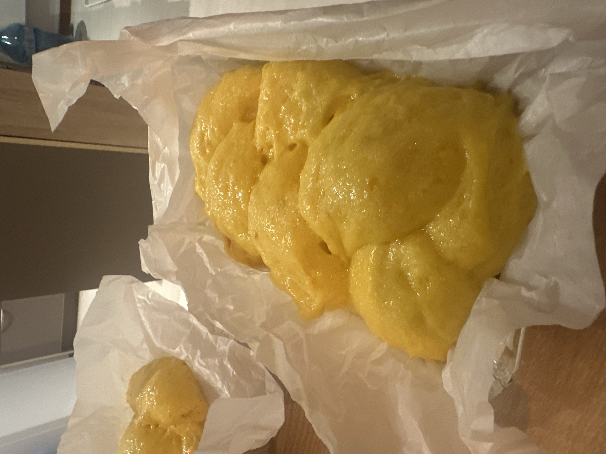 A close-up, high-angle shot depicts a portion of freshly made food on a sheet of crumpled white paper. The food appears to be a soft, golden-yellow dough, possibly related to bread, pastry, or potato, exhibiting a wet, glistening surface. The paper is on a dark wooden surface. The scene is indoors, presumably in a domestic kitchen setting, as indicated by the presence of a kitchen counter and a glimpse of appliances in the blurred background. There is no readily discernable information to place this image in Sofia, Bulgaria. There are no people visible in the image.