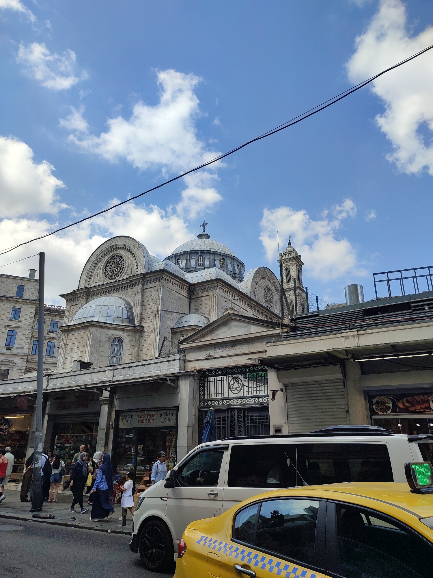 This is a street-level, medium-shot, eye-level, vertical photograph taken on a cloudy day in Beyoğlu, Turkey. The scene is dominated by the exterior of a grand stone building with domes and a bell tower, characteristic of Byzantine architecture. The building is partially obscured by a row of shops with awnings and signage, some of which display Turkish text. In the foreground, two vehicles are visible: a white van and a yellow taxi. People are walking on the sidewalk in front of the shops, and some are interacting. The sky is a vibrant blue, with scattered white clouds, and a dark power line stretches across the upper portion of the frame. The overall impression is that of a busy urban environment with historical and commercial elements coexisting.