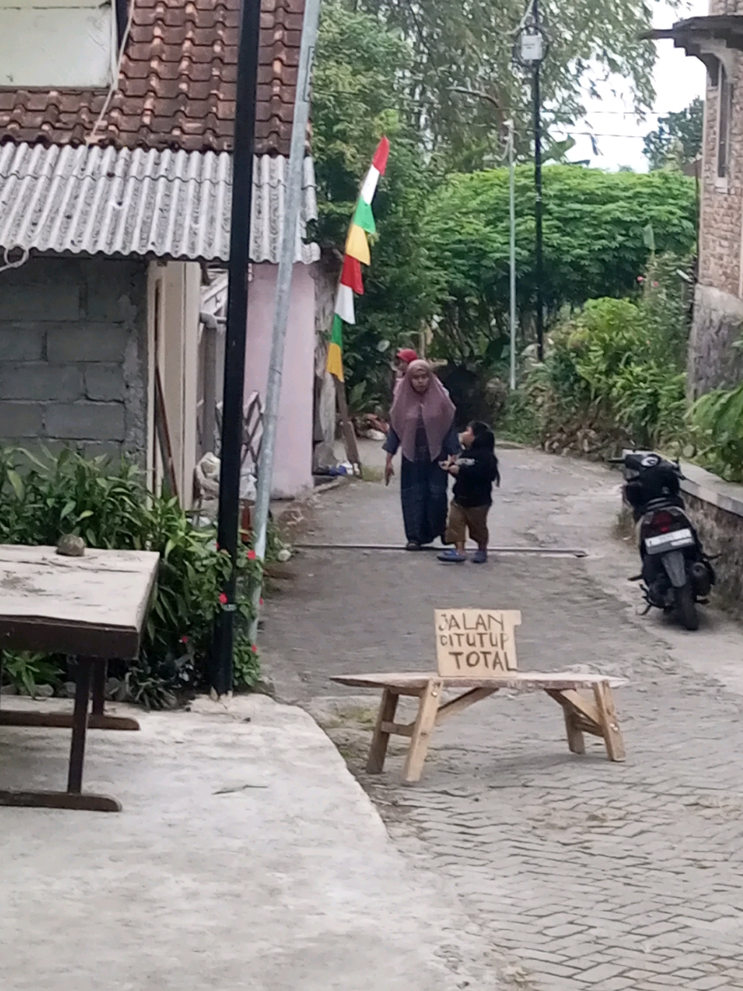 In Andongsili, Indonesia, a scene unfolds on a paved street lined with greenery and buildings. A woman in a hijab and a child are walking away from the camera. In the foreground, a small wooden table displays a sign that reads "JALAN DITUTUP TOTAL," indicating the road is completely closed. A motorcycle is parked on the right side of the street. The environment suggests a residential area with elements of Indonesian architecture visible in the background.