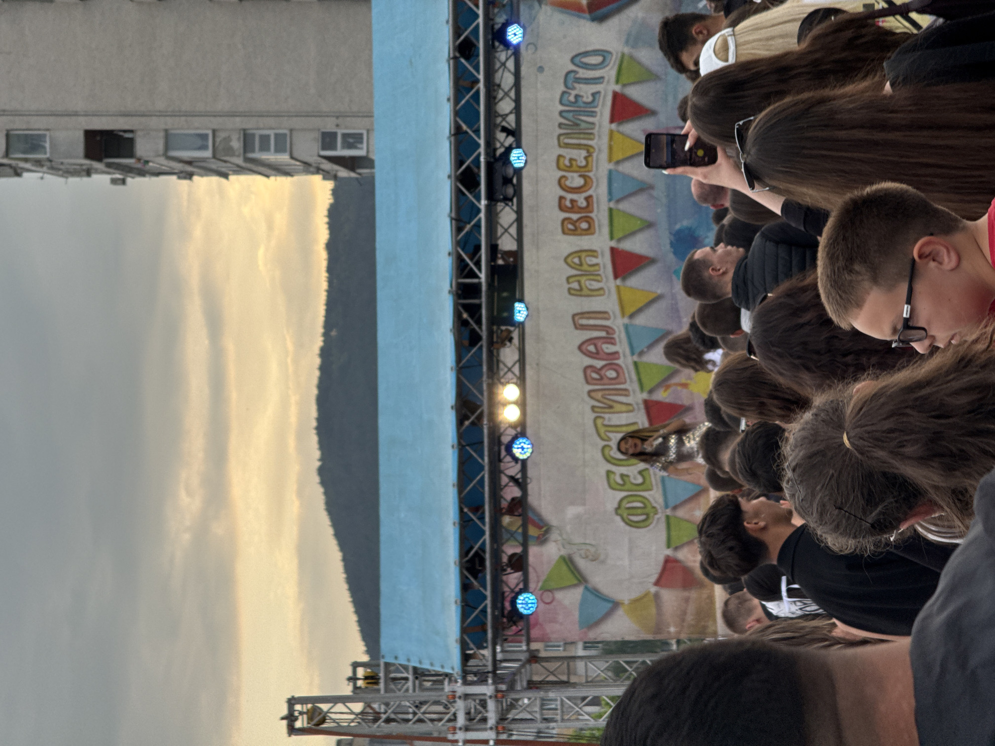 An outdoor festival or concert scene unfolds under an evening sky, with soft golden light visible through clouds. The perspective is from behind a crowd of attendees, looking towards a stage on the left. Many individuals in the foreground and mid-ground have dark hair, and some are holding up smartphones to record or photograph the event. One person's phone clearly shows a screen capture.
On the stage, illuminated by blue and white stage lights, a performer—a woman in a sparkly outfit—is visible. Behind her, a large backdrop displays colorful geometric patterns, resembling bunting, and features prominent text in Cyrillic script: "ФЕСТИВАЛ НА ВЕСЕЛИЕТО," which translates to "FESTIVAL OF MERRIMENT."
In the background, to the far left of the stage, a multi-story building with several windows is visible. Further in the distance, a dark, tree-covered mountain or hill stretches across the horizon under the fading light. The overall atmosphere suggests an engaged audience enjoying an outdoor performance as dusk approaches in Shumen, Bulgaria.