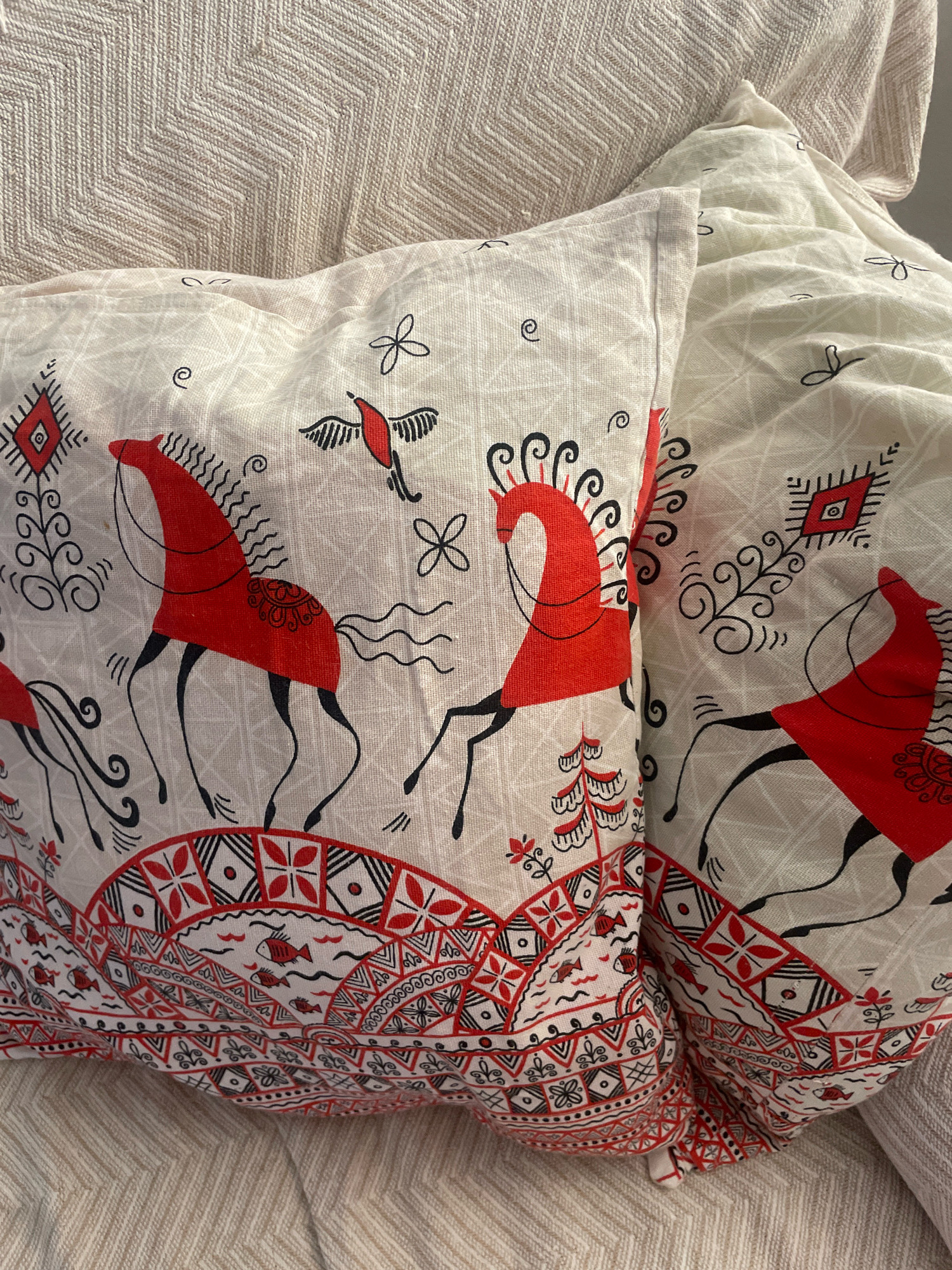 The image shows two pillows with a white background and a folk art design featuring red and black illustrations of horses, birds, trees, and geometric patterns. The pillows are placed on a textured beige fabric, possibly a sofa or bed. The lighting suggests an indoor setting, likely a domestic environment. The image does not provide any information about the city of Atarfe, Spain, or its location within the city.