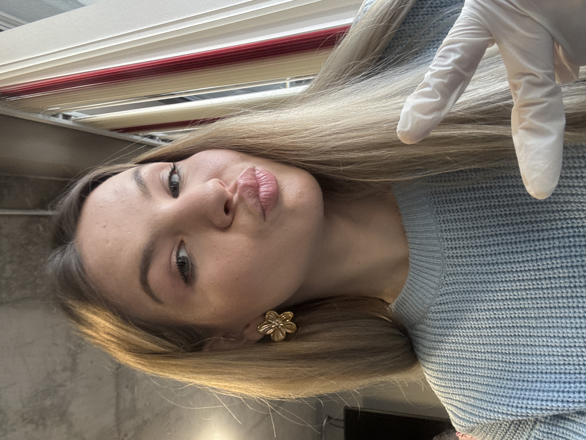 A selfie photograph captures a woman with light skin, blonde hair, and a light blue sweater. She is making a kissing face toward the camera. Her left hand is raised and wearing a white latex glove. She has a flower-shaped earring on her left ear. The background shows a set of horizontal blinds, suggesting an indoor location. The location is not discernible within the city of Plovdiv, Bulgaria from the image alone.