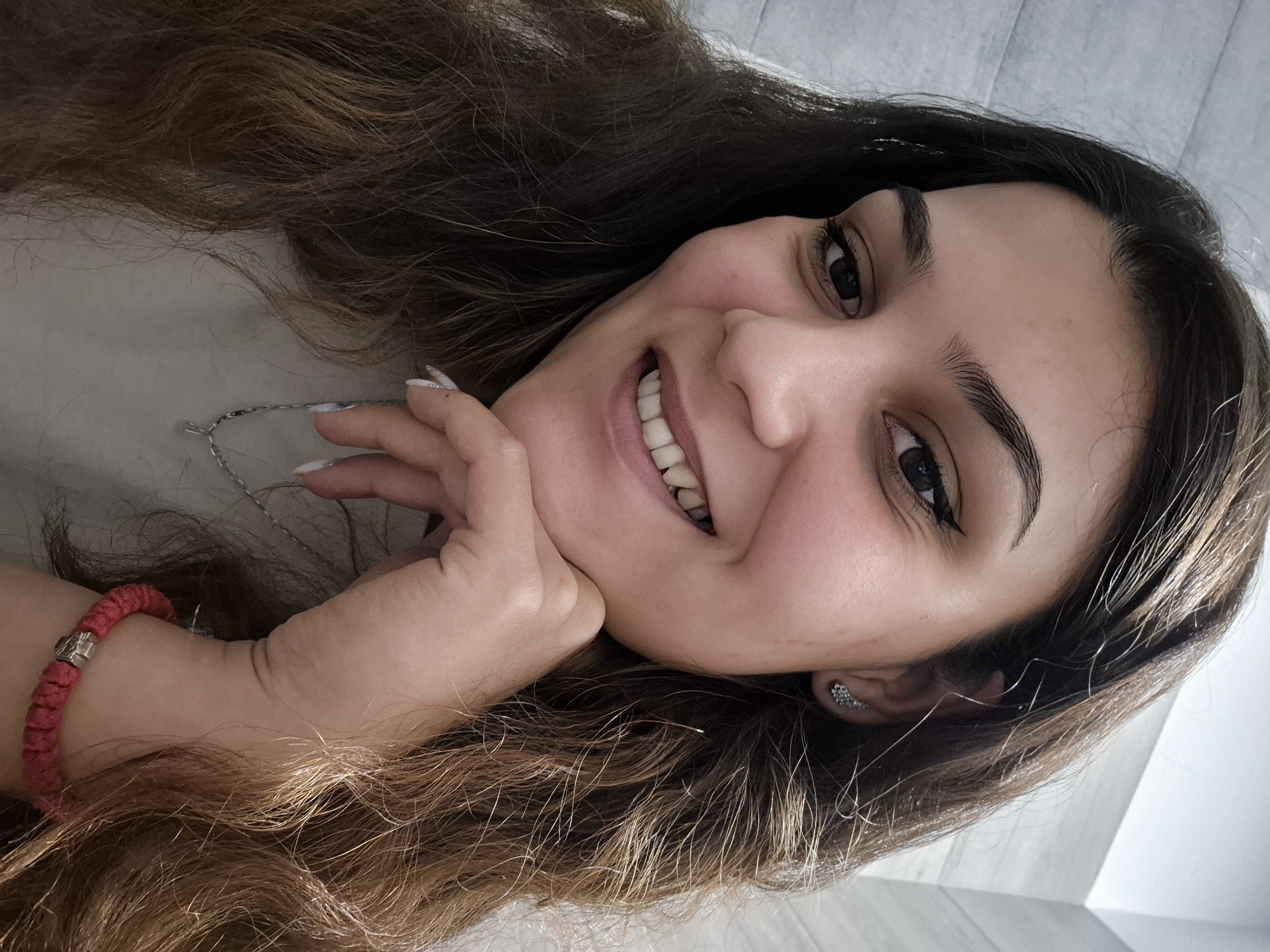 A close-up, brightly lit portrait captures a young woman smiling warmly, her head tilted slightly as she rests her chin on her left hand. She has fair skin, dark, expressive eyes with subtle eyeliner, and dark, well-defined eyebrows. Her wide smile reveals her teeth, conveying a sense of happiness and contentment.
Her long, wavy dark brown hair, with lighter brown highlights catching the light, cascades around her face and shoulders. She wears a delicate silver-colored chain necklace and a vibrant red braided bracelet with a silver clasp on her left wrist. A small, sparkling silver earring is visible in her right ear. Her fingernails are manicured with a light, possibly white or silver, polish.
The background suggests an indoor setting, with light-colored, subtly textured fabric behind her head, possibly a bed or sofa, and a lighter, smoother surface to her right, likely a wall or another piece of furniture. The soft, natural lighting suggests the photograph was taken during the daytime. The image appears to be a casual, personal portrait. No text is visible in the media file. The context provided indicates the location is Sofia, Bulgaria.