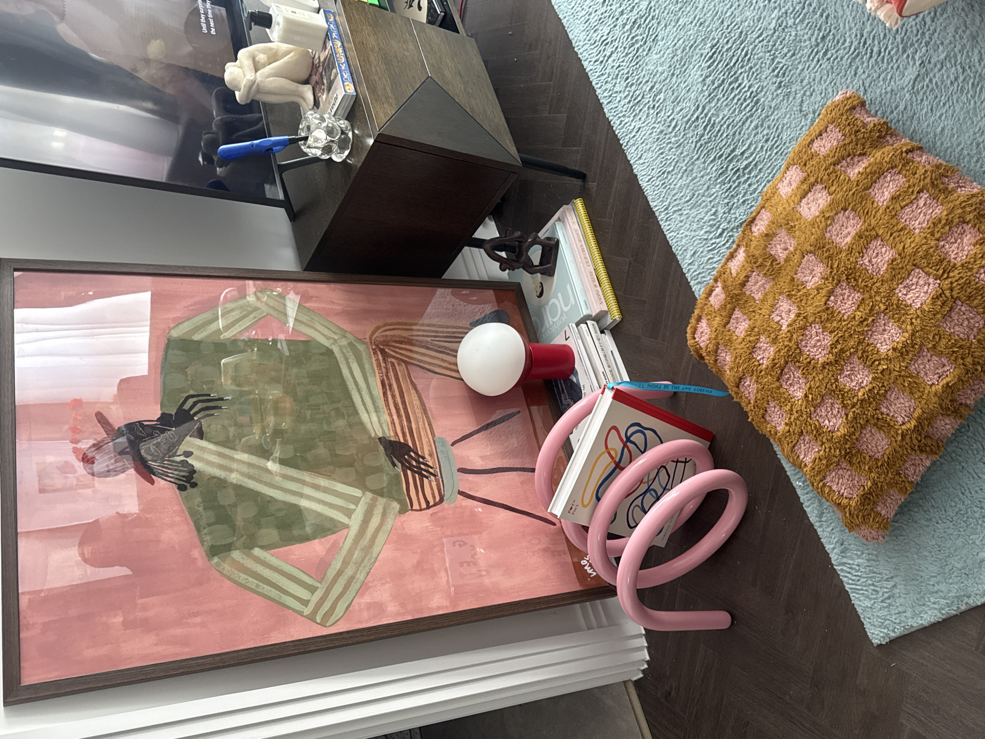 The image depicts a living room interior. A framed artwork hangs on the wall, featuring an abstract figure in shades of pink and green. The painting is partially obscured by various objects. Adjacent to the artwork, a dark brown cabinet houses a small decorative figure and a lighter. Several books, including one with the title "nar" visible, are stacked near the artwork, along with a white spherical lamp with a red base. A pink, coil-shaped object rests near the books. A checkered pillow in shades of brown and pink sits on a light blue rug. The rug lies on a dark brown wooden floor. A TV is visible above the cabinet.