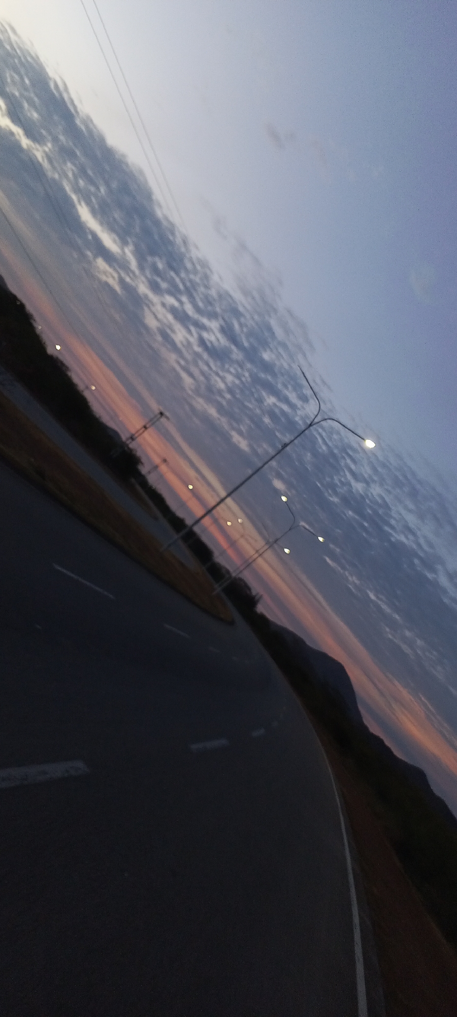 The image depicts a road with white lane markings, curving to the right and leading towards a distant mountain range. Streetlights are visible along the roadside. The sky shows a gradient of colors suggesting sunset or twilight, with clouds. The location is likely near Altagracia, Venezuela.