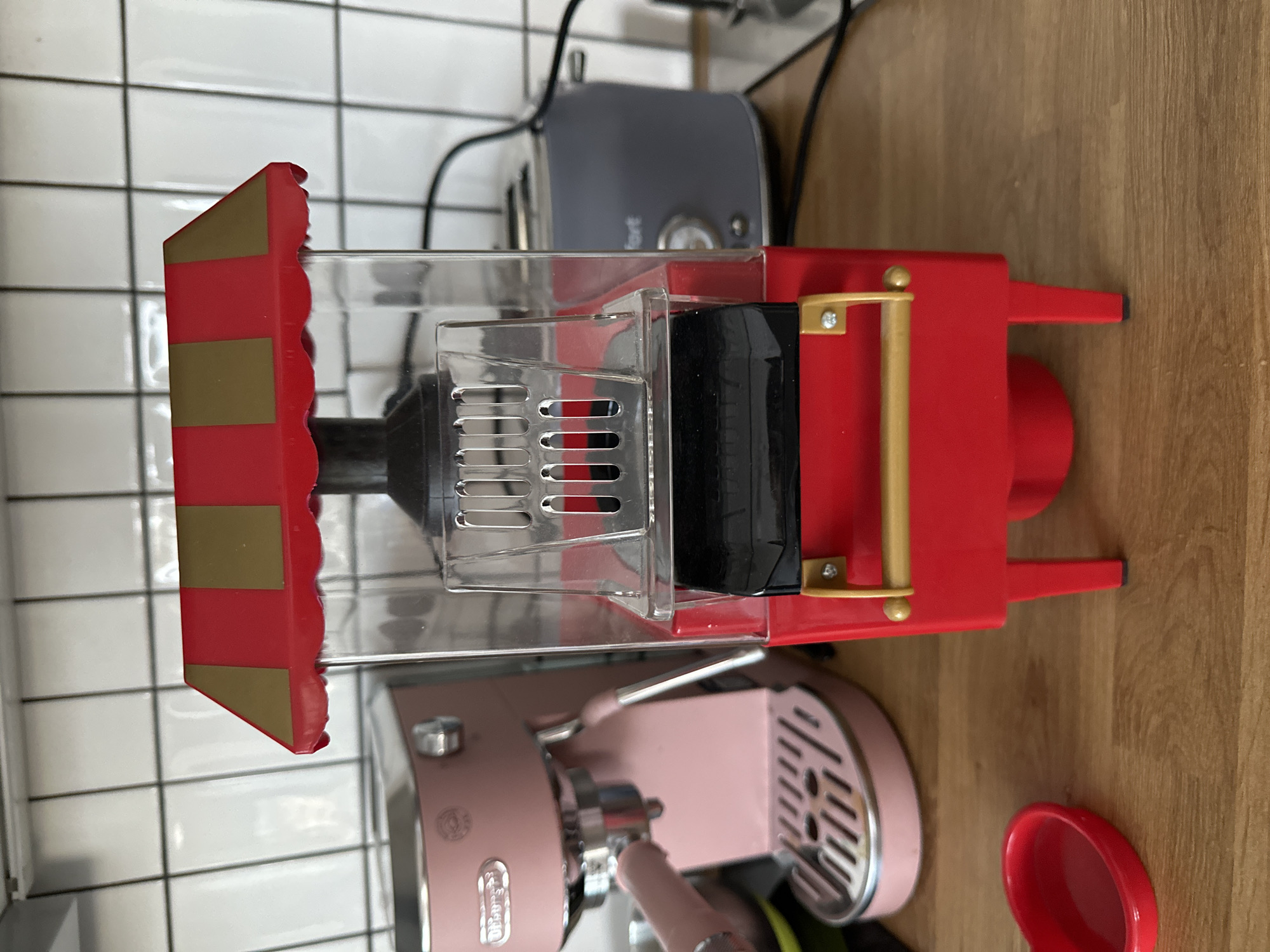 A red and gold striped popcorn machine sits on a wooden countertop in front of a tiled backsplash. To the left of the popcorn machine is a pink espresso machine, and to the right is a gray toaster. A red heart-shaped dish is on the countertop to the left of the popcorn machine. The scene is indoors, likely in a kitchen. There are no people visible in the image. The location is Vsevolozhsk, Russia.