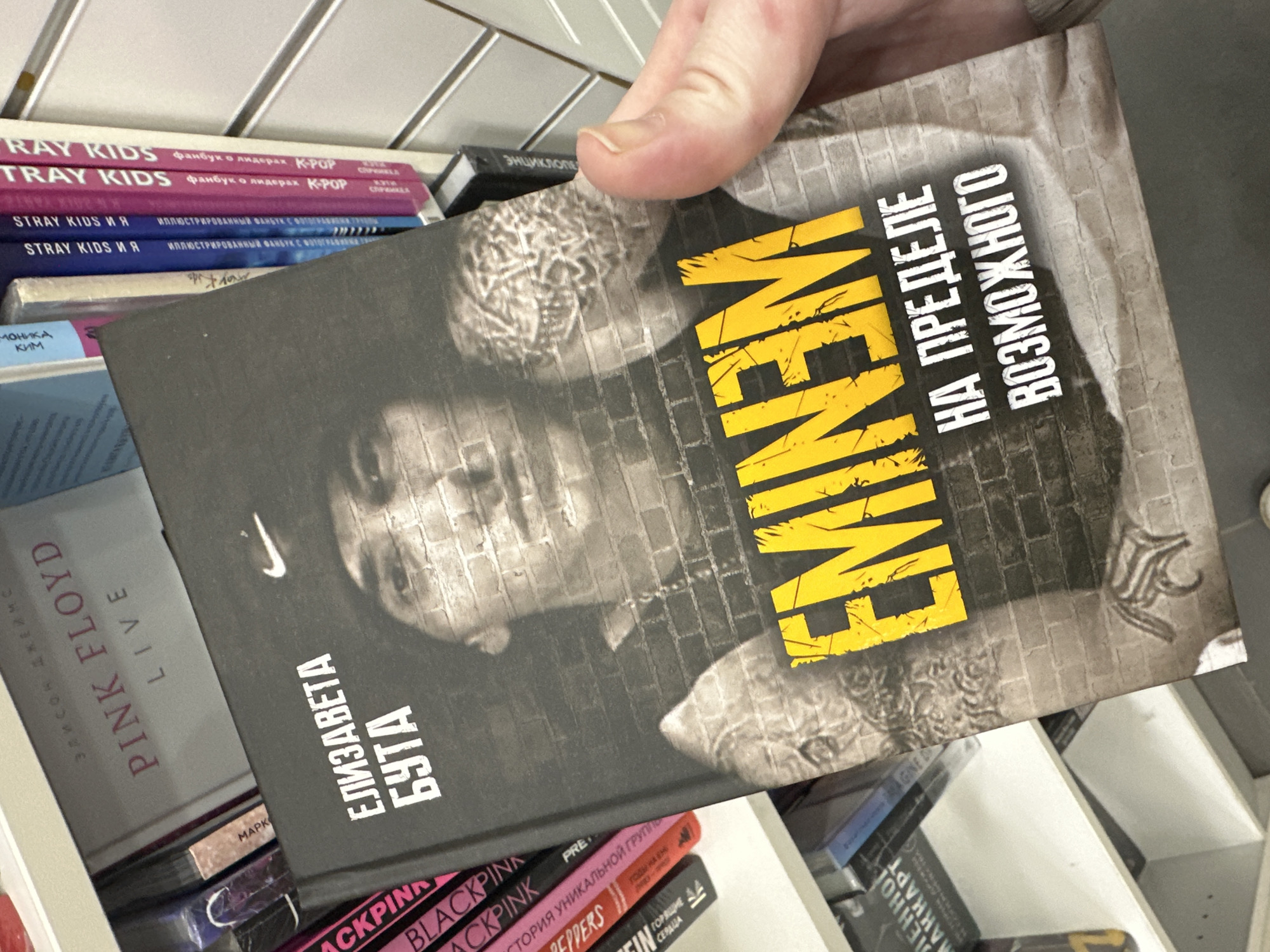 An eye-level shot captures a person's hand holding a book in what appears to be a bookstore or library in Izhevsk, Russia. The main subject is a biography featuring the rapper Eminem, held vertically by the top corner. The book's cover is dark grey with a brick wall pattern, and features a stylized, slightly faded image of Eminem's face. The title "EMINEM" is prominently displayed in large, distressed yellow capital letters. Below it, in white text, are the Russian words "НА ПРЕДЕЛЕ ВОЗМОЖНОГО" (On the Limit of the Possible) and "ЕЛИЗАВЕТА БУТА" (Elizaveta Buta), likely the author. A faint Nike swoosh logo is also visible on the cover.
In the background, shelves are filled with other books, predominantly fan books and biographies related to music artists and K-Pop groups, indicating a specific section of the store. Visible titles include "STRAY KIDS" and "STRAY KIDS И Я" (Stray Kids and I), with descriptions like "Фанбук о лидерах K-POP" (Fanbook about K-POP leaders) and "ИЛЛЮСТРИРОВАННЫЙ ФАНБУК С ФОТОГРАФИЯМИ" (Illustrated Fanbook with Photos). Other books include "PINK FLOYD LIVE" by "ЭЛИСОН ДЖЕЙМС" (Alison James), "BLACKPINK" with "СТОРИЯ УНИКАЛЬНОЙ ГРУППЫ" (History of a Unique Group), and partially visible "PEPPER" (likely Red Hot Chili Peppers), and "МОНИКА КИМ" (Monica Kim). The books are in various colors, including pink, purple, blue, and black. The lighting suggests an indoor setting.