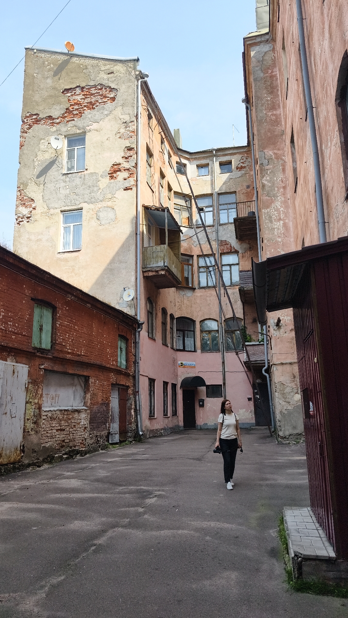 A young woman with dark hair, wearing a light-colored t-shirt, black pants, and white sneakers, walks away from the camera in a narrow, cobblestone courtyard. She carries a black camera and sunglasses in her right hand. The courtyard is surrounded by old, weathered buildings with peeling paint and exposed brick. To the left, a red brick building has large, metal-paneled doors and green-shuttered windows. To the right, a narrow, dark red wooden structure with metal detailing runs along the side of the courtyard. The buildings in the background are a pale pink and beige, with multiple windows, balconies, and exposed pipes. The sky above is clear and blue. The scene appears to be in Vyborg, Russia, suggested by the architecture and the sign on the pink building which reads "Комната" (Room in Russian).