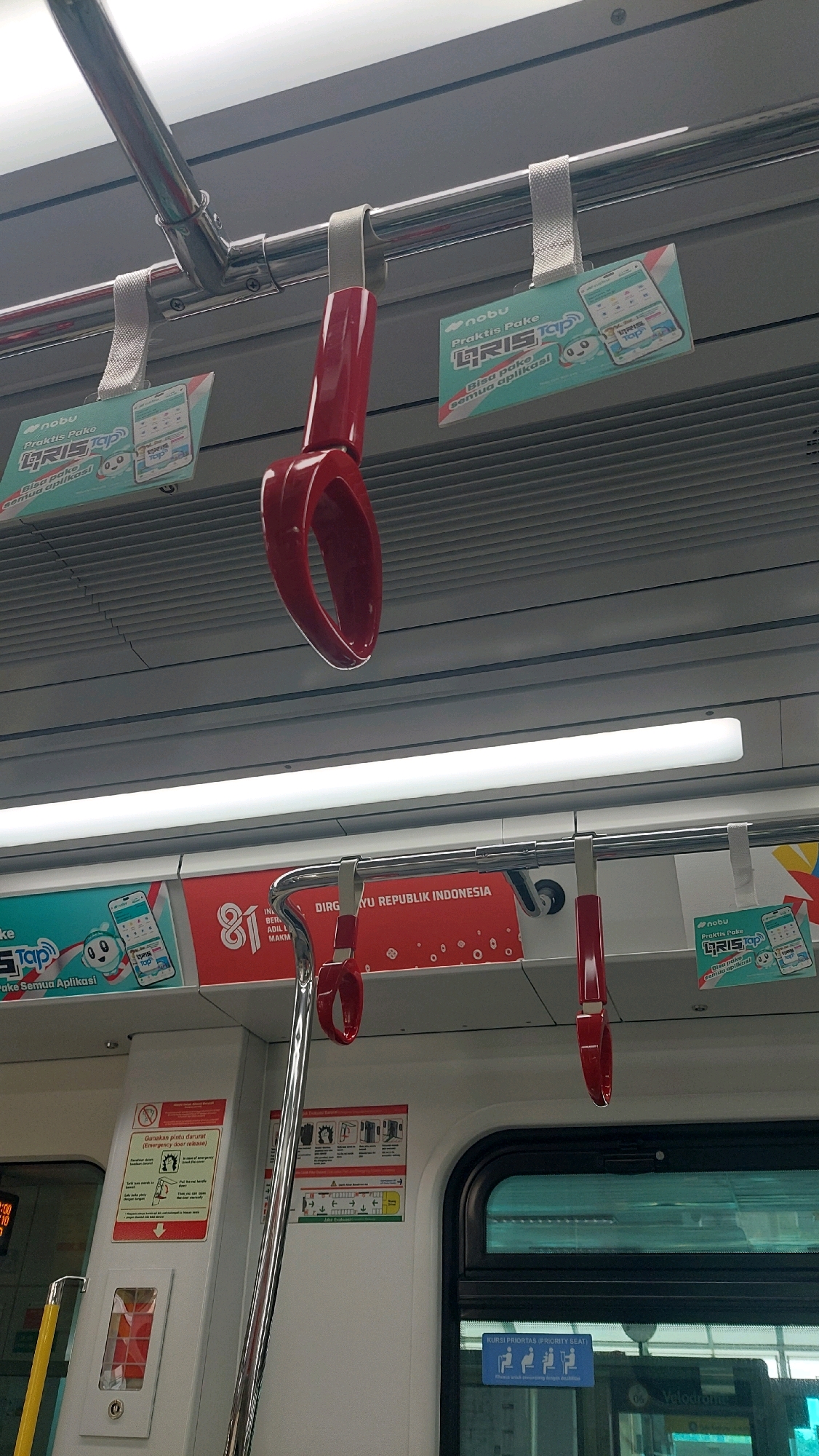 The image depicts the interior of a train carriage, likely a light rail or commuter train, in East Jakarta, Indonesia. The focus is on the overhead handrails and hanging straps, characteristic of public transport. Several advertisements are visible, primarily for a mobile application called "Nobu GRIS TOP," featuring a turquoise and white color scheme. One large advertisement on a red background commemorates "81 INDONESIA." A prominent, bright white fluorescent light runs horizontally across the upper portion of the frame, illuminating the scene. The handrails are made of polished metal, and the hanging straps and handles are a vibrant red. To the lower left, a yellow pole is partially visible, and a white panel with red text and diagrams, possibly related to emergency procedures, is mounted on the wall. A blue sign indicating "KURSI PRIORITAS (PRIORITY SEAT)" with icons of seated individuals is visible on the right side. The overall environment is clean and functional, typical of modern urban public transportation.