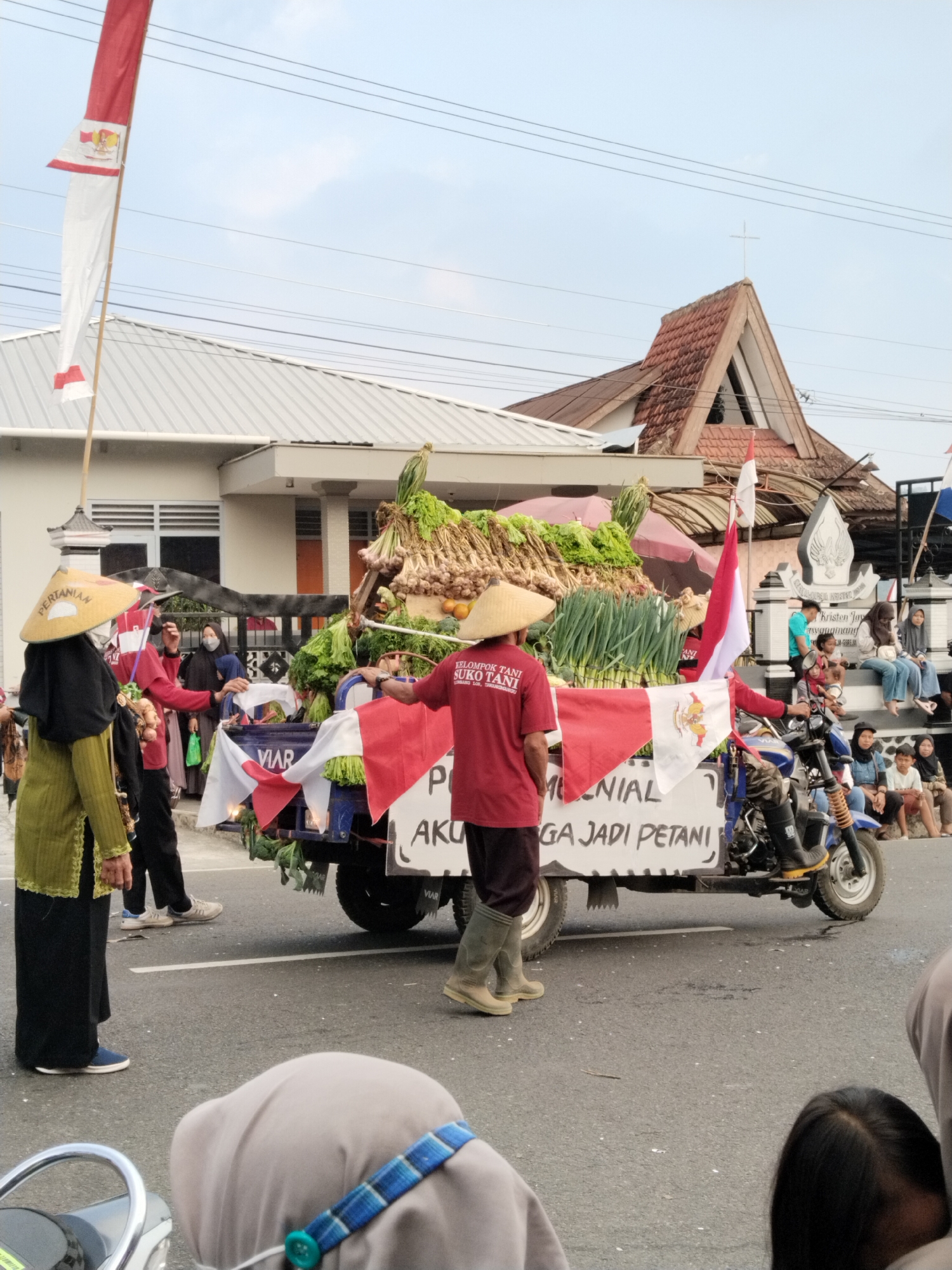 The image depicts a parade or procession in Nglebak, Indonesia. In the foreground, a person wearing a green tunic, black trousers, and a conical straw hat stands on the left. To their right, another person in a red shirt and tall rubber boots faces away from the camera, standing beside a cart loaded with produce. The cart is decorated with Indonesian flags and a sign that reads "MELONIAL AKU JADI PETANI." Various vegetables, including lettuce, onions, and other leafy greens, are prominently displayed on the cart.
A motor vehicle, likely a trike or scooter, is attached to the rear of the cart and appears to be powering the procession. Several other individuals, some wearing headscarves and masks, are visible in the background, either participating in the parade or observing from the roadside. Buildings, including a house with a white roof and a building with a red tiled, steeply pitched roof topped with a cross, line the street. The sky is overcast. The overall scene suggests a community event celebrating agriculture and local farmers.