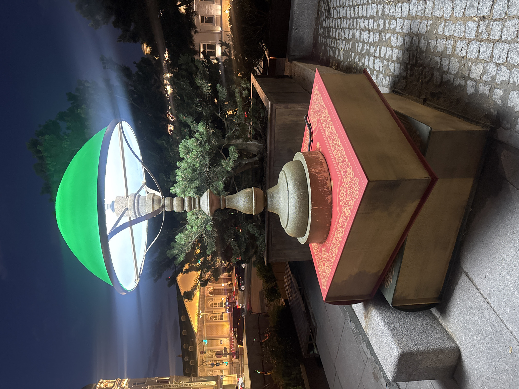 An outdoor decorative lamp with a bright green shade is situated on top of two large, oversized books. The top book is red with gold decorative patterns on its cover, and the bottom book is green. The lamp itself has a decorative metal base. The scene is set at dusk or night, with a dark sky and artificial lighting illuminating the background. In the background, there's a building with illuminated windows, and a street with cars and traffic lights. The lamp and book sculpture is placed on a cobblestone ground next to a stone planter with greenery. This installation is located in Moscow, Russia, specifically near the Bolshoi Theatre on Teatralnaya Square. There are no people visible in the image.