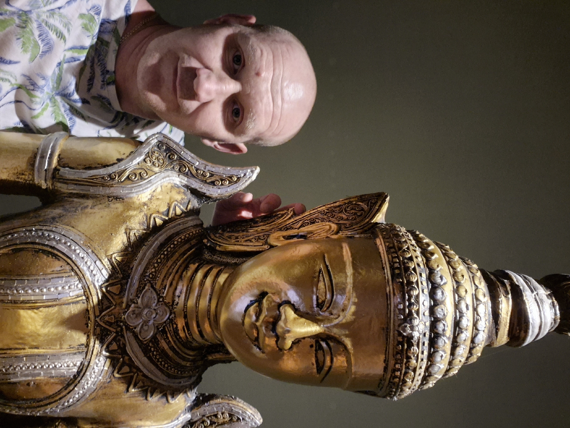 A bald man in a floral shirt looks down at a golden Buddha statue. The statue is highly ornate, with layers of decoration. The man's hand is partially touching the statue. The background is a solid dark color. The location and its associated city, Alanya, Turkey, cannot be confirmed from this image alone.