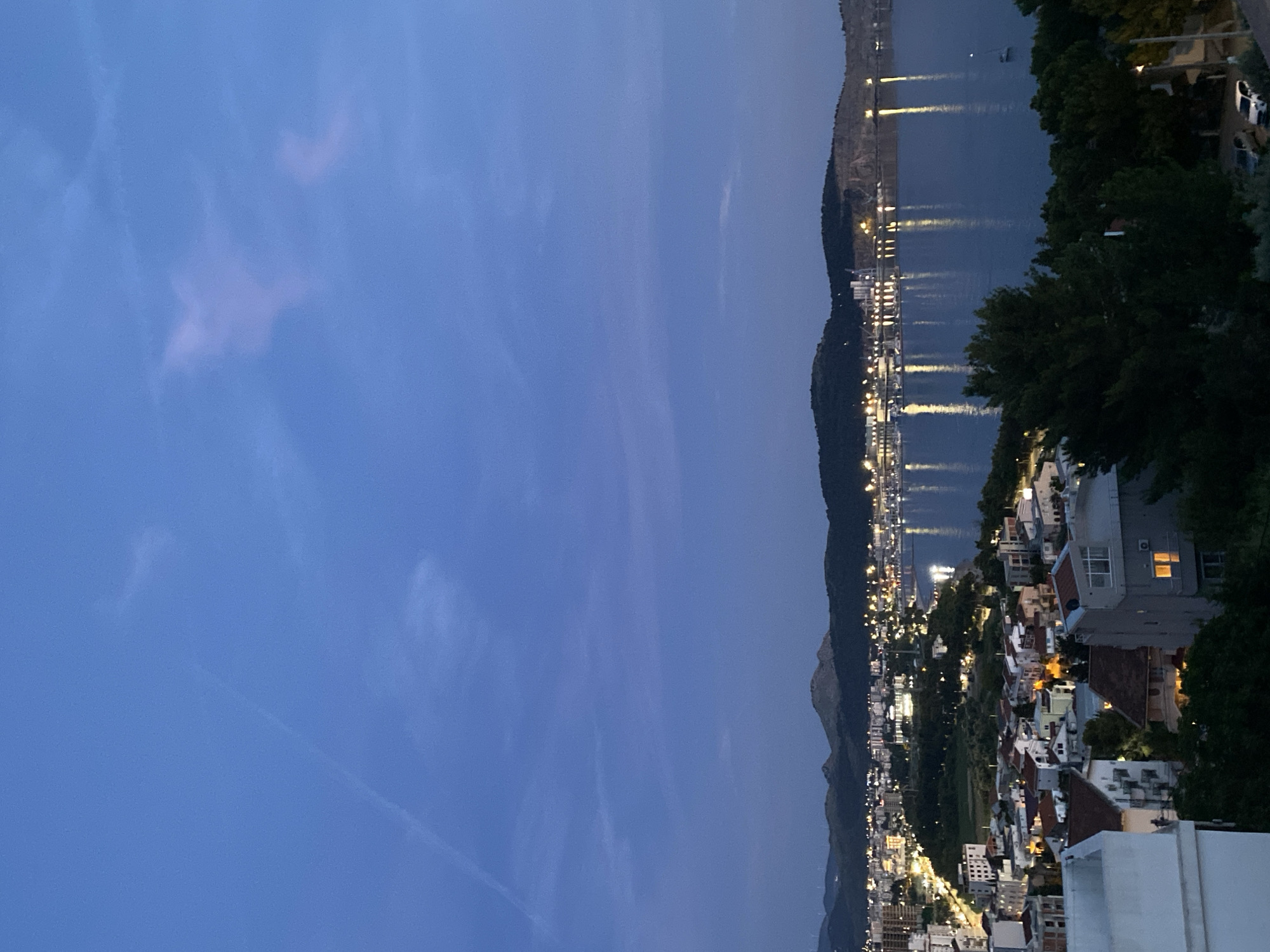 An aerial, high-angle, wide shot captures the city of Šušanj, Montenegro, at dusk. The sky, a deep twilight blue, features wispy clouds and a faint contrail. The city sprawls along the coast, a dense collection of buildings with lit windows and streetlights casting a warm glow. Buildings range from multi-story apartment blocks to smaller, red-roofed houses. In the foreground, partially visible white and gray buildings are silhouetted against the darkening landscape. Lush green trees fill the lower right portion of the frame. Beyond the city, a wide expanse of calm water reflects the lights from the shore and scattered boats, creating shimmering vertical streaks. In the distance, dark, rolling hills form the backdrop, their peaks dotted with more lights. The overall scene conveys a peaceful transition from day to night.