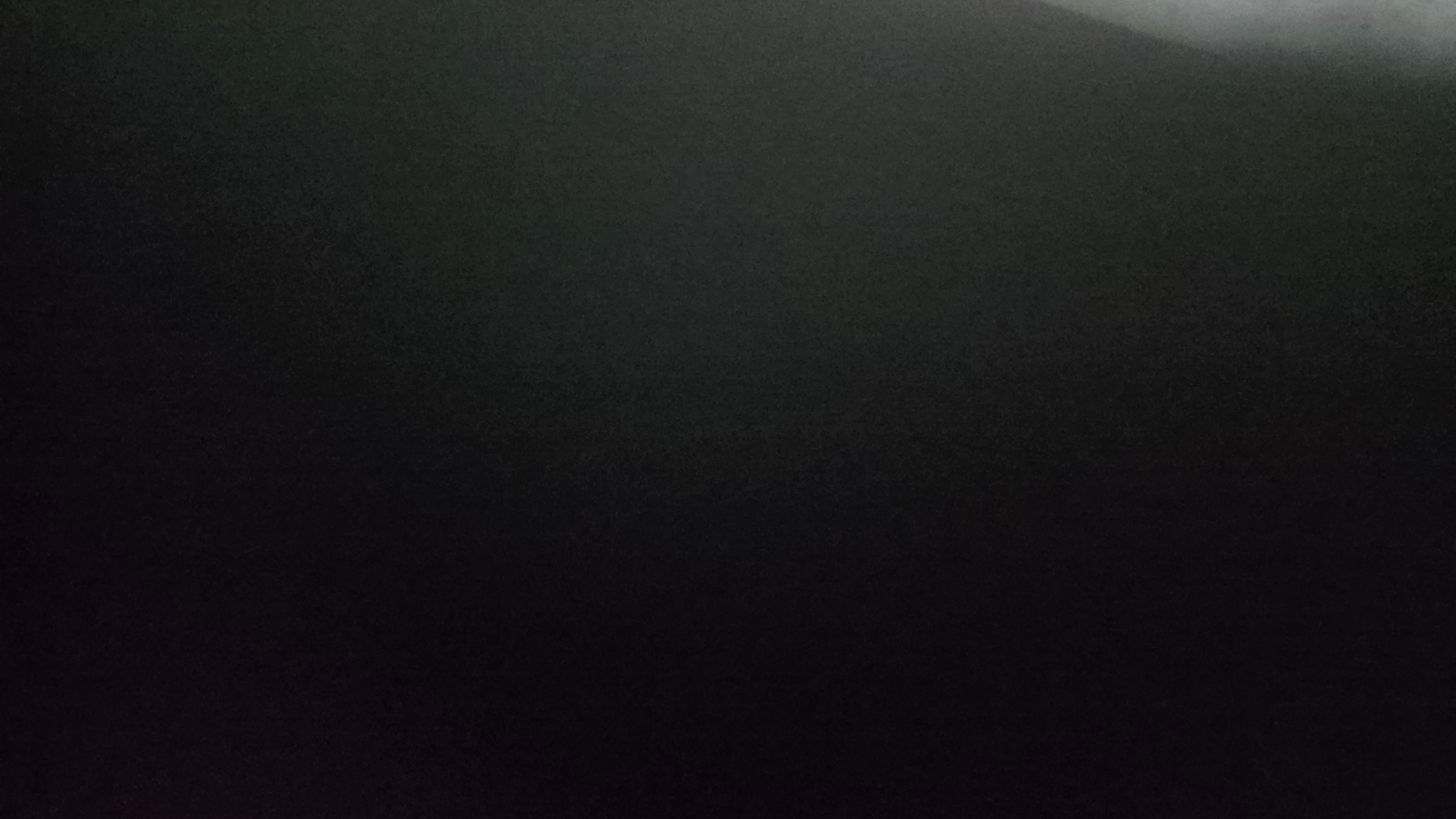 The image is overwhelmingly dark, primarily consisting of very deep gray and black tones. A consistent, fine grain or digital noise is distributed throughout, giving a slightly textured appearance to the otherwise undifferentiated darkness.
Subtle color variations are present: a faint, indistinct reddish-purple hue is visible in the upper-left quadrant, and a delicate greenish tint appears in the upper-right and central-right portions. Along the extreme right edge, an irregular vertical band of light gray to white is observed, suggesting a highly reflective surface or an external light source partially captured at the frame's periphery. Its form is indistinct, preventing identification.
No discernible subjects, objects, people, actions, or specific environmental features can be identified within the frame. The image provides no visual information to determine a specific geographical location, including the city of Dobrich, Bulgaria.