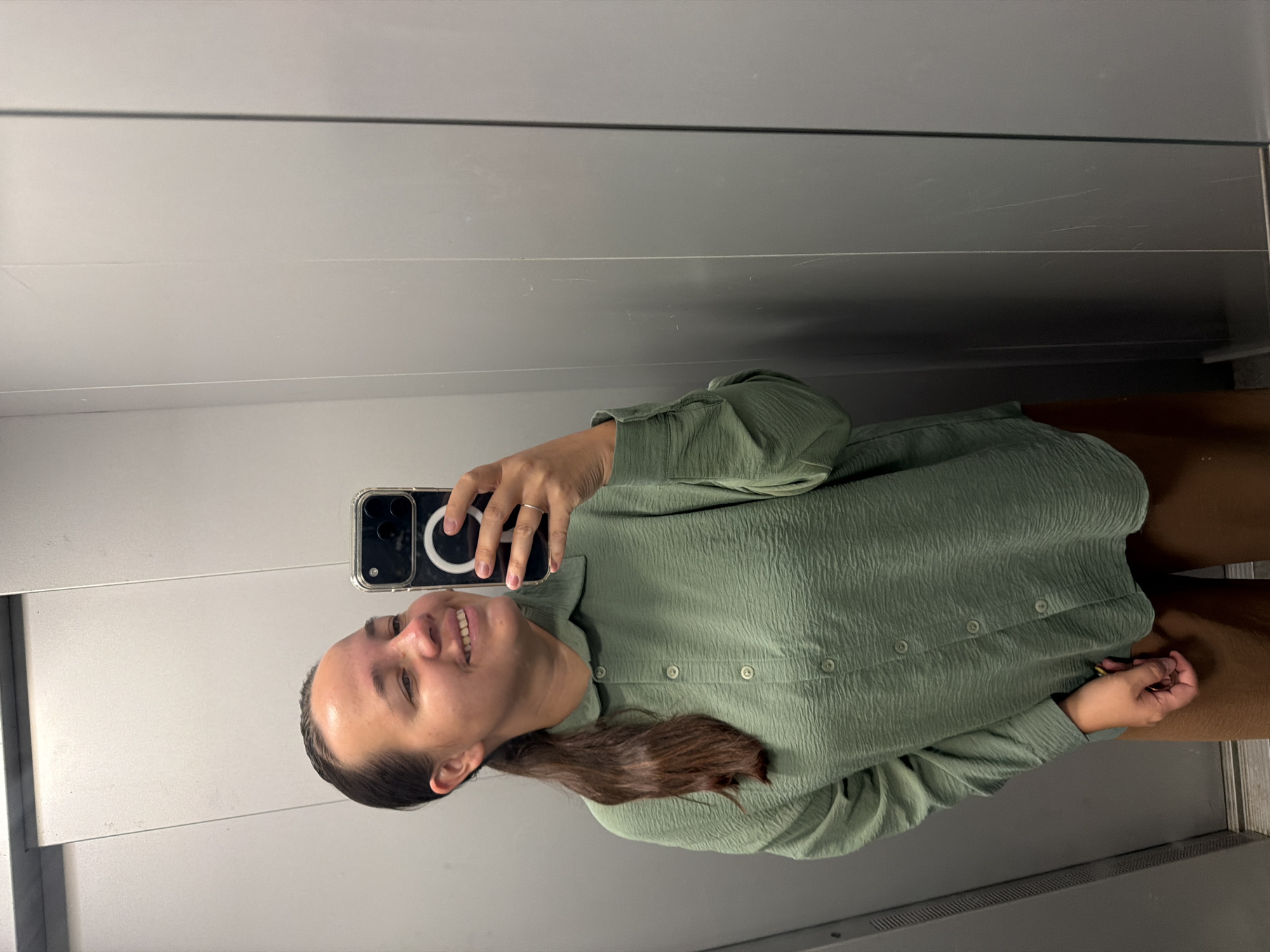 A young woman with dark hair pulled back in a ponytail takes a selfie in an elevator. She is wearing a textured green button-down shirt and brown pants. The elevator walls are gray and appear to be made of metal panels. The overall lighting is dim. The image was taken in Chaikovsky, Russia.