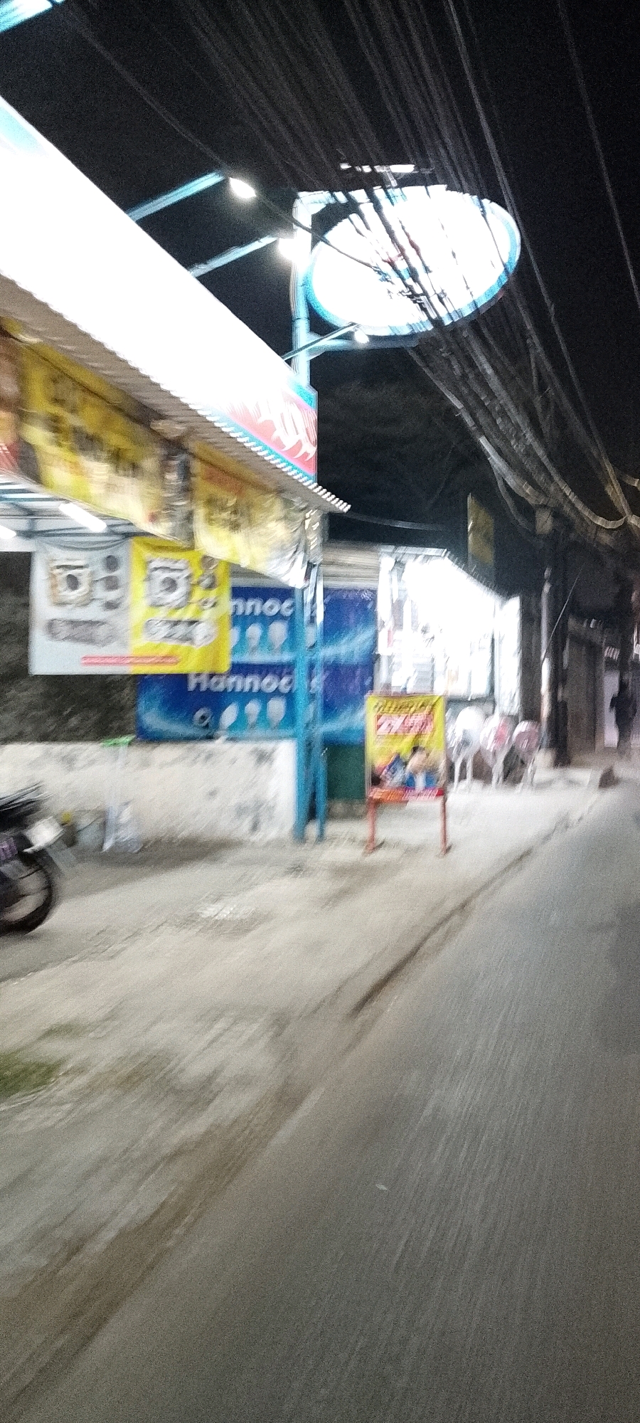 The image captures a street scene in Depok, Indonesia, at what appears to be night or dusk. The perspective is from a moving vehicle, indicated by the motion blur affecting most of the scene. On the left, a white awning covers a storefront. Below the awning, two signs are visible. The left sign is rectangular and appears to display illustrations of electronic devices, possibly cameras. The right sign is yellow and features text that is partially obscured but includes "Hannoc." Further to the right, a large blue banner with a repeating pattern of what look like light bulbs and the text "Hannoc" is displayed. A bright yellow sign on a red stand is positioned further down the street, with an image of people and text that is unreadable due to blur. A motorcycle is partially visible on the far left, also blurred by motion. Overhead, a dense network of electrical wires crisscrosses the scene. The road in the foreground is paved and appears wet in places. The background shows the dark silhouettes of buildings and trees. A few indistinct figures are present in the distance on the right side of the street.