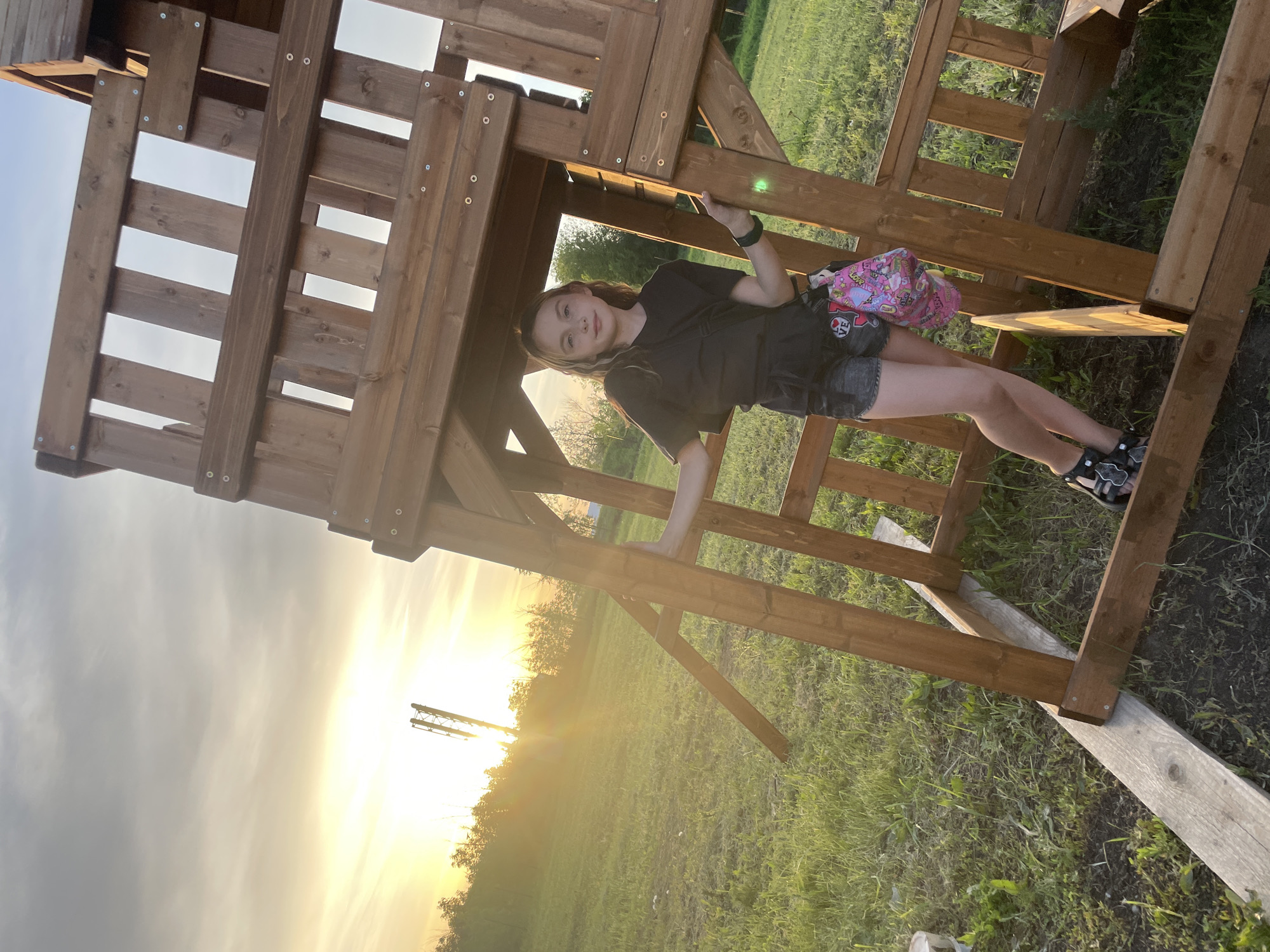 A young person is climbing on a wooden play structure in an outdoor setting. The structure appears to be a playhouse or fort, with wooden beams and supports forming the framework. The individual has on a dark-colored shirt, shorts, and sandals. They are holding onto the structure with their hands, and a pink backpack rests on their side. The environment consists of grassy areas, trees, and a vibrant sunset visible in the background. The precise location is not identifiable as Saratov, Russia from the image.