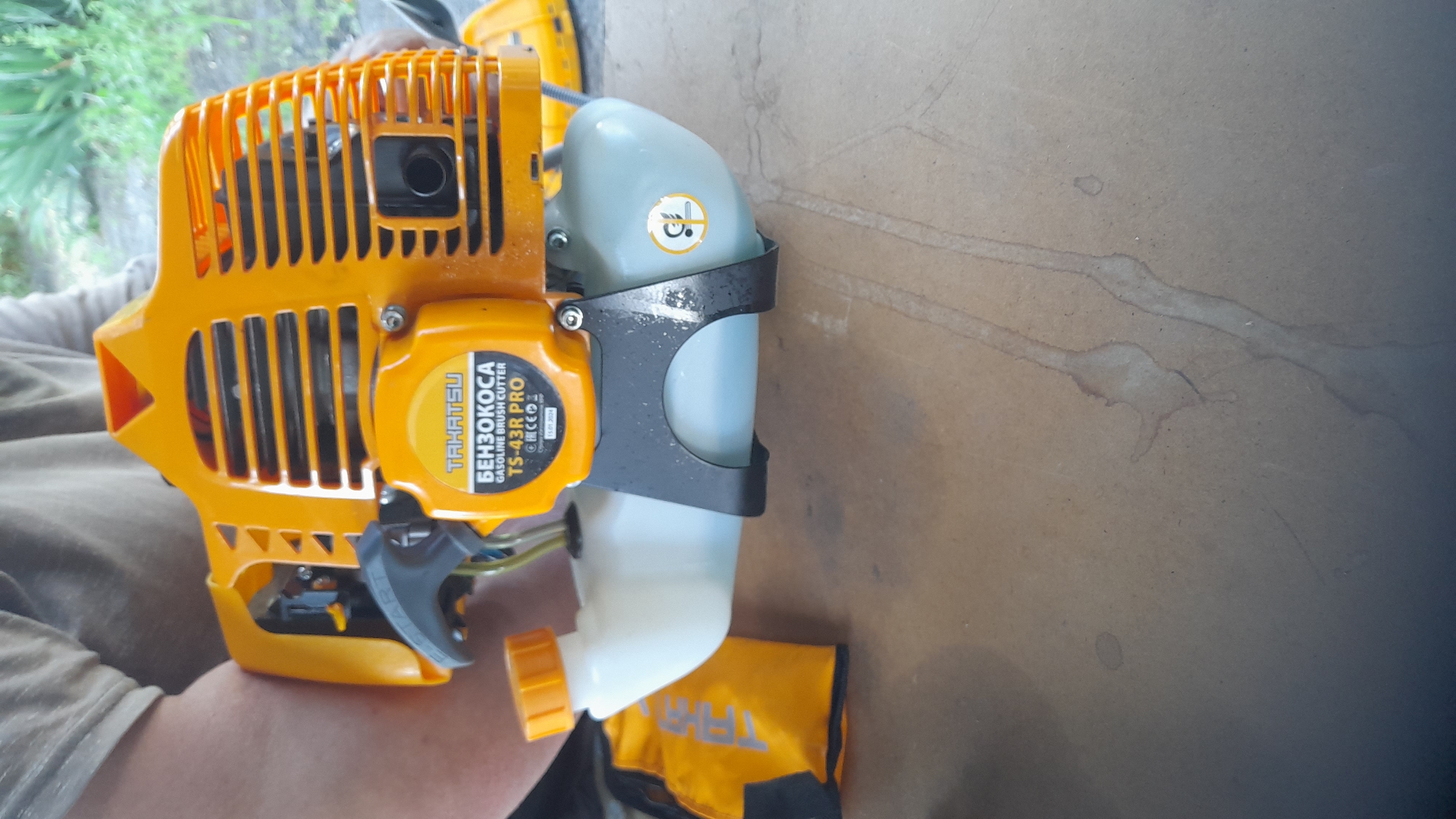 A top-down, medium shot captures a person in a light-colored shirt holding a bright orange gasoline brush cutter. The brush cutter is the primary subject, with its fuel tank, engine housing, and control handle clearly visible. A yellow fabric accessory, possibly a carrying bag or cover, is positioned to the left of the brush cutter. The background shows a blurred natural setting with green foliage, suggesting an outdoor environment. The brush cutter is placed on a textured, light brown surface, which appears to be a table or workbench. A label on the brush cutter reads "TAHATSU BENZOKOSA GASOLINE BRUSH CUTTER TS-43R PRO." A circular yellow sticker with a graphic is also visible on the fuel tank. The lighting is diffused, and the overall image has a slightly muted color palette. The scene appears to be taking place in or around Bagayevskaya, Russia, likely in a residential or workshop setting where such equipment would be stored or maintained.