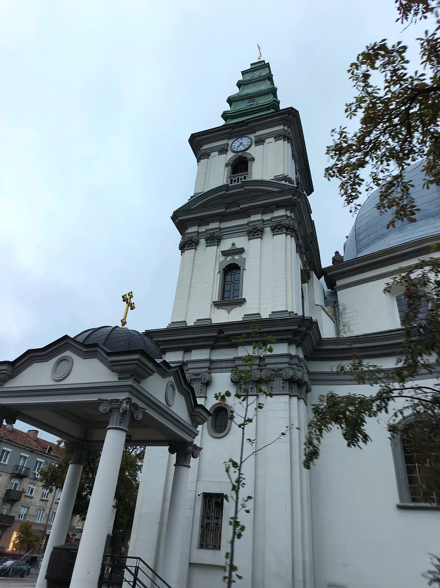 The image shows a low-angle view of the exterior of the Ternopil Transfiguration Cathedral in Ternopil, Ukraine. The cathedral is a white, neo-baroque building with a tall, green, tiered bell tower featuring a clock. A golden cross is visible at the apex of the bell tower. The main structure of the cathedral has grey decorative elements and columns. In the foreground, a portico with large white columns supports a grey roof. To the left of the portico, an apartment building with balconies is visible. The sky is overcast. Branches of trees with green and reddish leaves partially obscure the view.