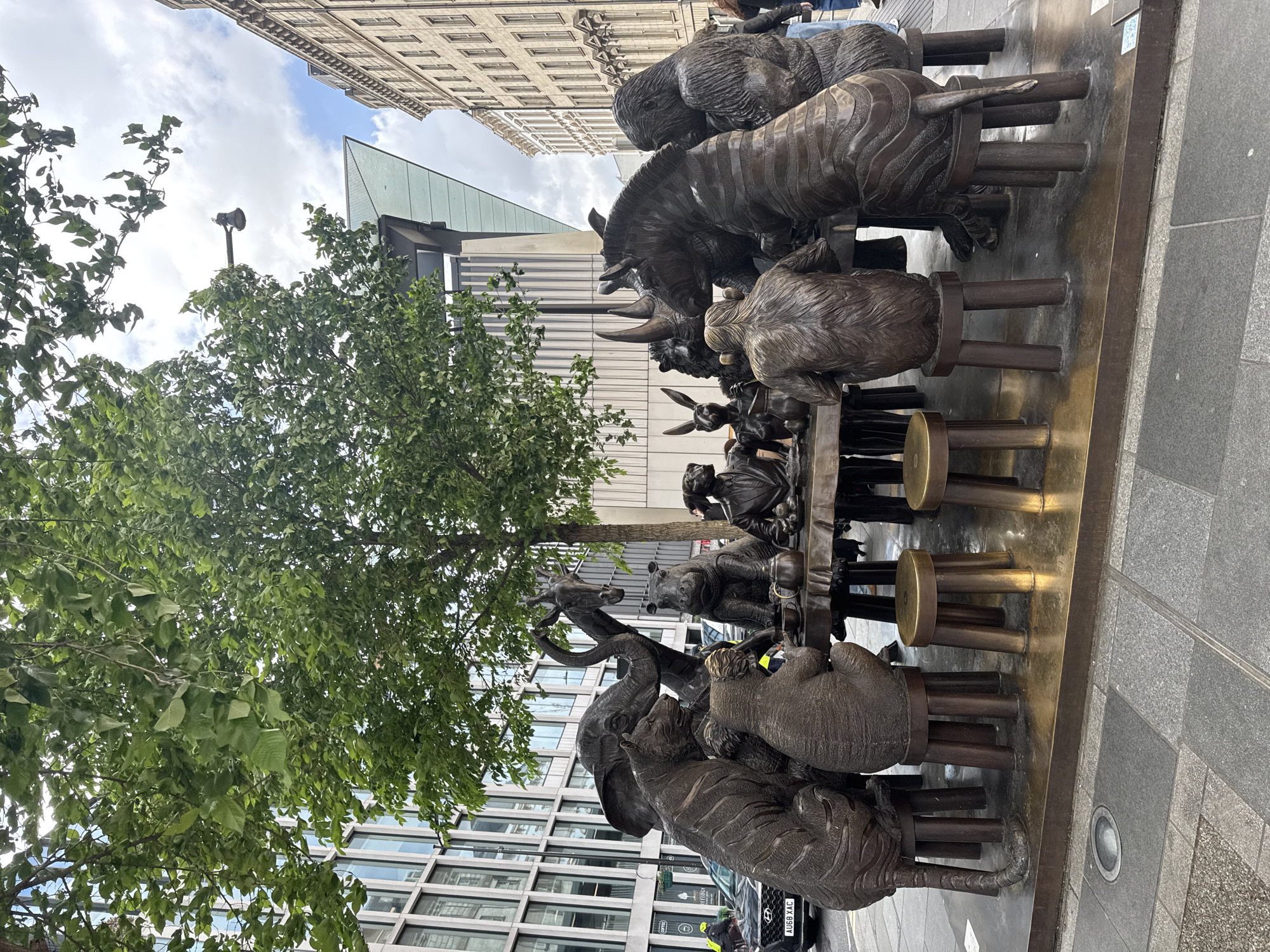 Here is a description of the image:
The image depicts a bronze sculpture of animals seated around a table. Animals include a tiger, elephant, giraffe, rhinoceros, bear, zebra, gorillas, monkeys, and rabbits. The sculpture is situated on a city street with buildings in the background. A tree with green leaves is present to the left of the sculpture. The location is an unknown city. A black car, its license plate marked 'AU68 XAC', is visible.