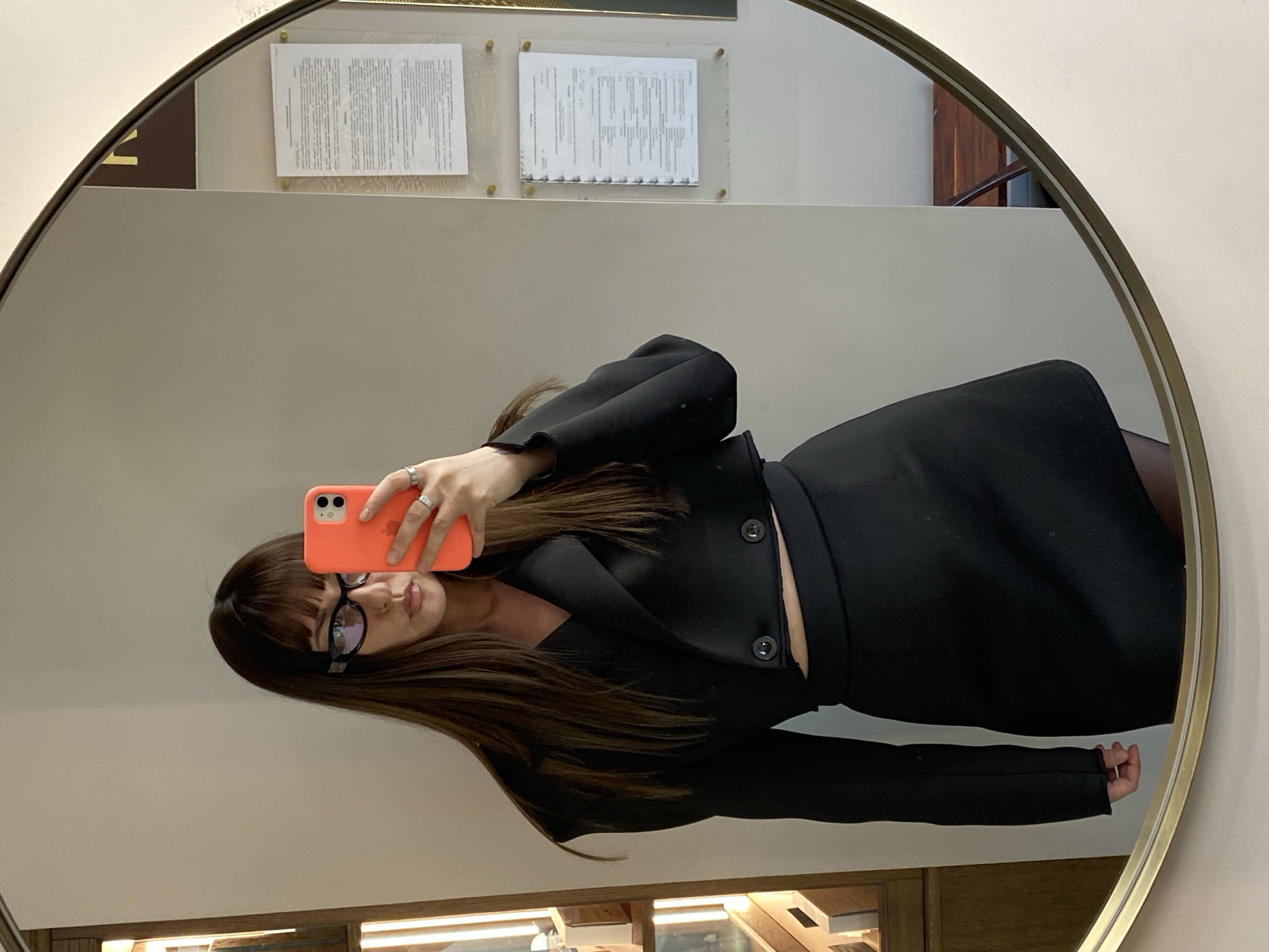 A person is captured in a mirror selfie, reflected horizontally within a round, gold-framed mirror. The individual, appearing female, has long, dark brown hair with bangs, wears black-rimmed glasses, and holds a smartphone with a vibrant orange case in her left hand (as reflected). She is dressed in a professional-looking black blazer-style top with buttons and a matching black skirt or dress, adorned with several rings on her left hand. Her expression is neutral and composed.
The background reflected in the mirror reveals a light-colored wall displaying two documents in transparent frames or holders. The text on these documents is clearly in Cyrillic script, consistent with the specified location context of Votkinsk, Russia, suggesting they are official forms or regulations. Below the documents, a segment of wooden furniture or a counter is partially visible. The indoor setting features uniform artificial lighting, providing no discernible details regarding the time of day or outdoor weather.