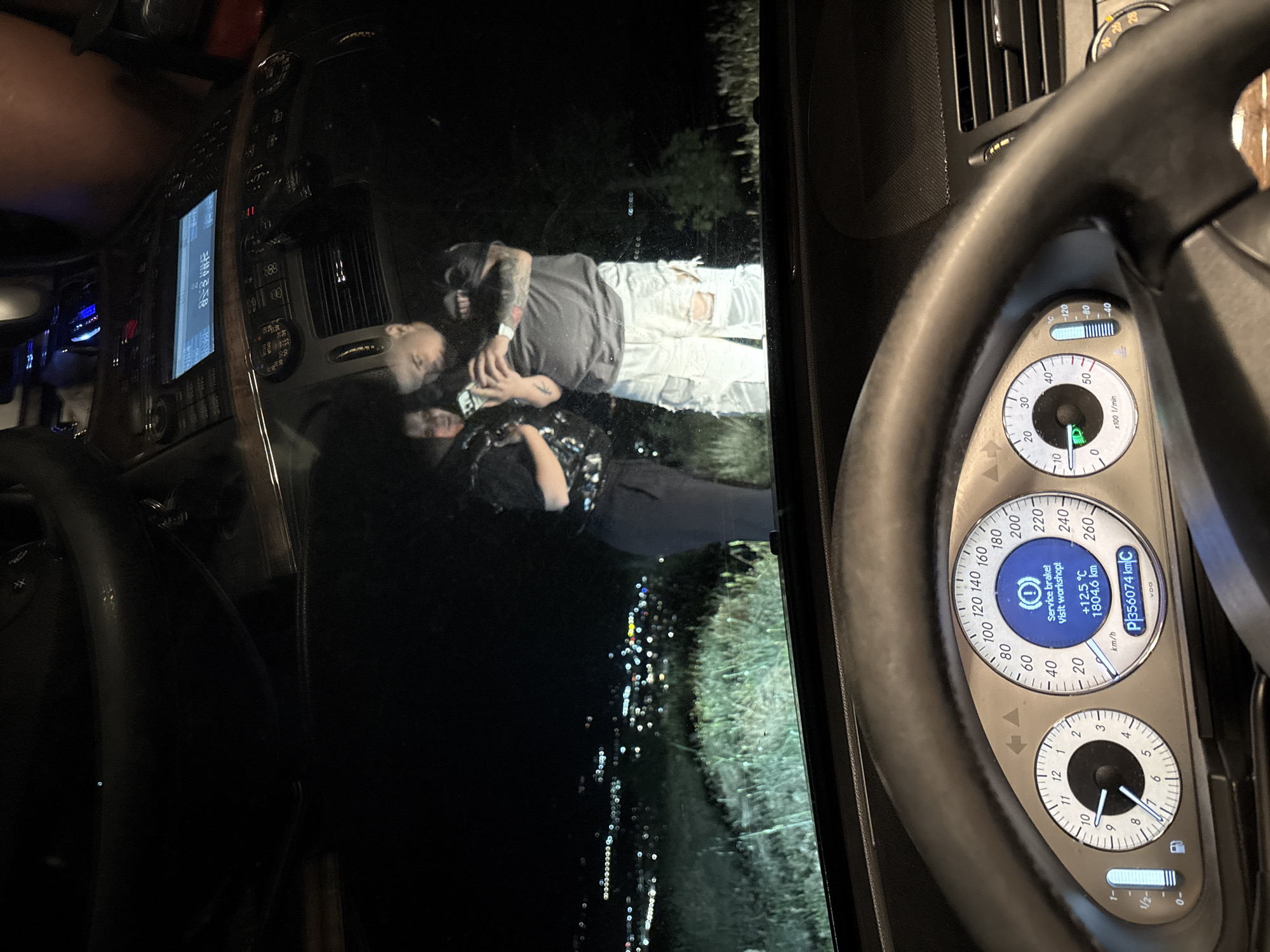 A view from inside a vehicle showing the dashboard and steering wheel. The speedometer reads a distance of 1804.6 km with a brake system service message illuminated. The temperature display reads 12.5°C. Outside the vehicle window, an image, possibly of a mobile phone, shows two people lying on grass at night, with city lights visible in the background. The location of Marulevo, Bulgaria is not indicated in the scene, and cannot be assumed.