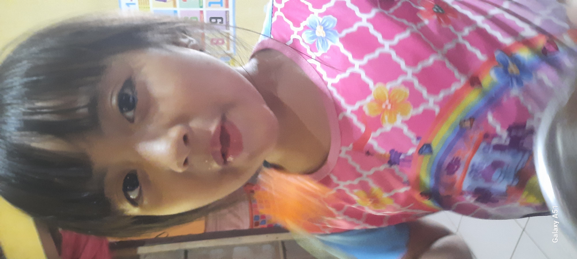 This is a vertical, close-up, candid photograph of a young Indonesian child, likely a girl, in an indoor setting. The child is the primary subject and is positioned in the lower half of the frame, tilted slightly to the right. Her face is turned towards the camera, with dark hair framing her face. Her eyes are dark and open, and her mouth is slightly ajar, with pink lips. She appears to be around 3-5 years old.
She is wearing a bright pink t-shirt adorned with a white lattice pattern and colorful floral motifs, including a light blue and a yellow flower. A rainbow graphic with purple and blue elements is also visible on the right side of her shirt, along with what appears to be depictions of cartoon characters or mythical creatures in purple and blue. A glimpse of a bright blue garment can be seen underneath the pink shirt. To the right of the child, a portion of a metallic, silver-colored object, possibly a bowl or pot, is visible. In the background, there is a blurry yellow wall with what seems to be educational posters displaying letters and numbers in various colors, suggestive of a play area or educational space.
The lighting suggests an indoor environment, likely during the daytime, given the natural light illuminating the scene. There are no overt indications of the weather. The overall mood of the photograph is informal and spontaneous, capturing a moment with the child. The text "Galaxy A51" is visible vertically along the right edge of the image, indicating the phone model used for the photograph. The setting is identified as Jatitengah, Indonesia.