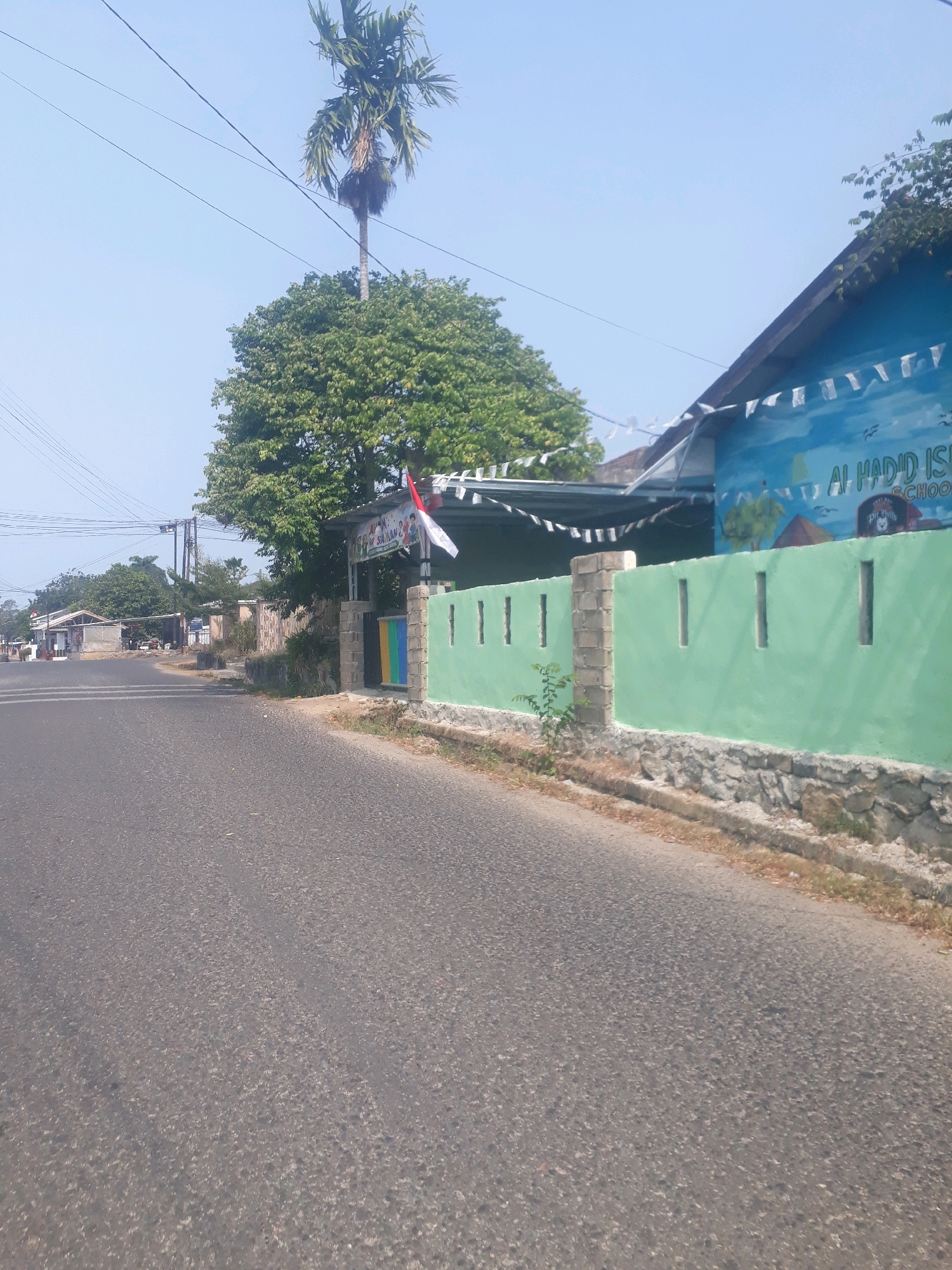 The image captures a street scene in Pangkalpinang, Indonesia, on a clear, sunny day. The foreground is dominated by an asphalt road, which curves slightly to the right. Along the right side of the road is a low, light green painted concrete wall topped with rough stone. This wall has rectangular cutouts at intervals. Behind the wall, a building with a blue facade is visible, featuring a mural. Text on the mural reads "AL HADID ISLAMIC SCHOOL". White triangular flags are strung along the roofline of the building. A tall palm tree stands to the left of the building, behind a large, leafy green tree. In the distance, to the left of the road, are several low-rise buildings and structures, suggesting a commercial or residential area. Power lines are visible across the sky. A small plant grows at the base of the green wall.