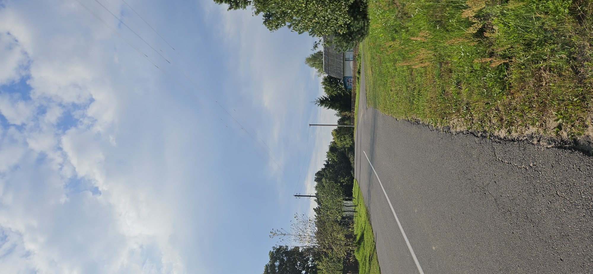 The image shows a rural road with a white line marking the center. The road curves to the right and is bordered by green grass and some dry stalks of grass. On the right side of the road, there is a small, light-colored house with a dark roof and blue trim around the windows, set back from the road and behind a wooden fence. The house appears to be in a rural area of Мазалаўскі сельскі Савет, Belarus. Trees line the background on both sides of the road, with more dense trees on the left and a mix of trees and a taller evergreen on the right, near the house. The sky is blue with scattered white clouds. Several power lines are visible stretching across the sky. There are utility poles along the side of the road. The road surface shows some cracking and wear. No people are visible in the image.