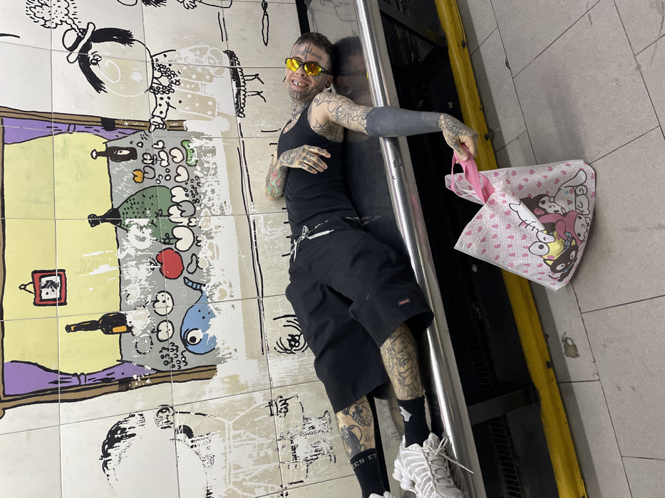 A tattooed man with yellow glasses and a black tank top is posing against a subway wall with cartoon artwork. He's wearing black shorts, black socks, and gray sneakers. His left arm is completely covered in black ink. He's holding a pink-handled bag with cartoon characters on it. The wall art features drawings of food, characters, and other items in a whimsical style. The scene appears to be on a subway platform with a silver railing behind the man.