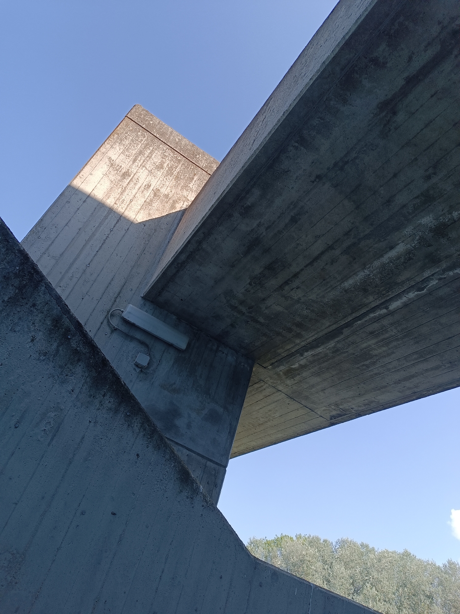 The image depicts a low-angle shot of a concrete structure, possibly part of a bridge or overpass, against a clear blue sky. Two large, angular concrete beams intersect in the frame. One beam, illuminated by sunlight, casts a shadow on another, darker beam. A light fixture with a gray casing is attached to the side of one of the concrete elements. In the distance, a line of trees with light-colored foliage is visible below the structure. The scene suggests an outdoor urban or suburban environment, likely in Casalmaggiore, Italy, given the context. There are no people or animals present in the image.