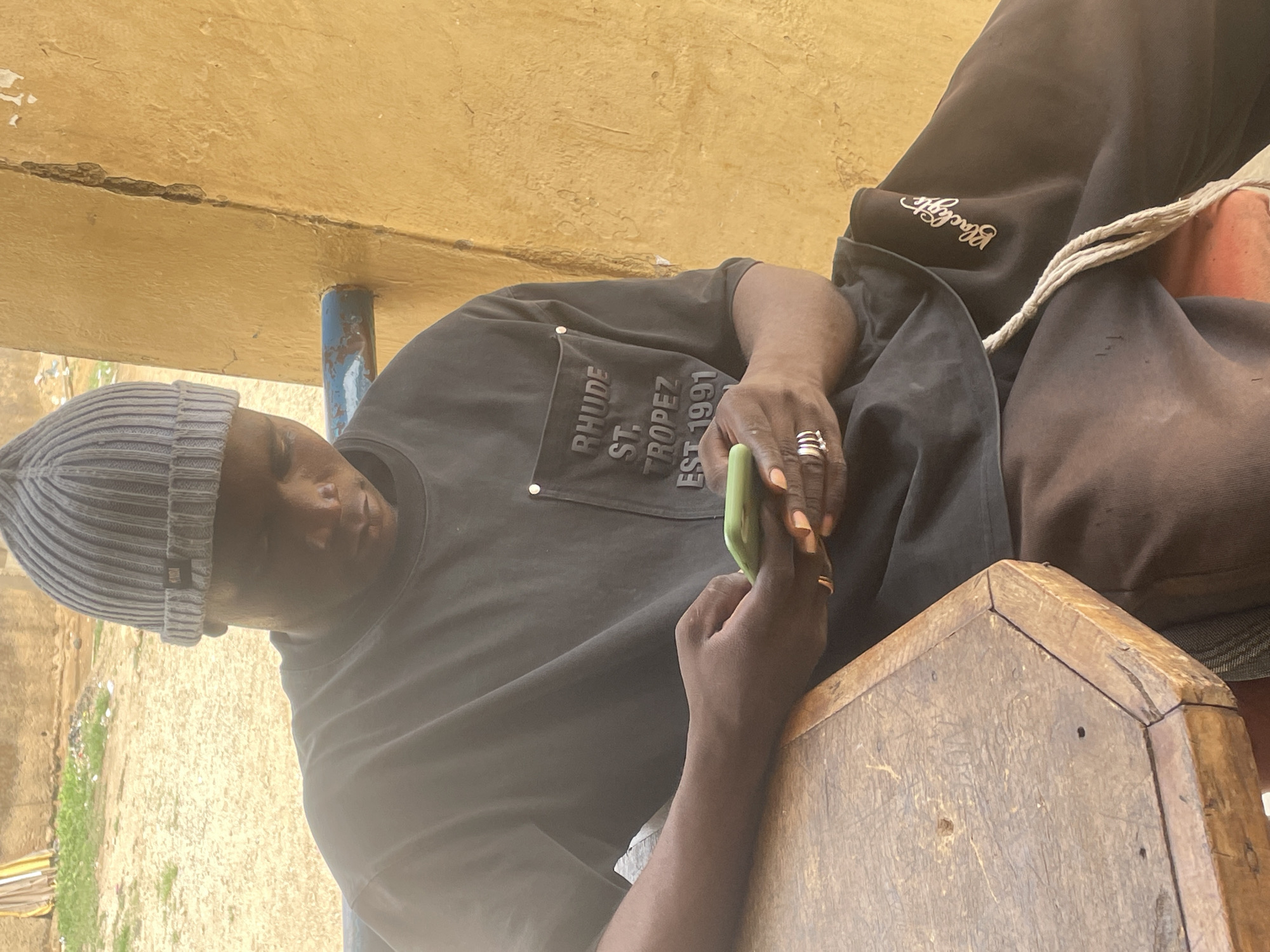 A man sits on a wooden stool in what appears to be an outdoor or semi-outdoor location in Jos, Nigeria. He is wearing a gray beanie, a dark t-shirt with the words "RHUDE ST. TROPEZ EST 1991" printed on the upper left side, and dark sweatpants or trousers. The sweatpants have the word "Blackgate" embroidered on the upper thigh. He is holding a green smartphone in his hands and appears to be looking at it. The stool he is sitting on is simple and constructed of weathered wood. There is a beam and support pole behind him, and some vegetation visible in the background. The location appears to be modest and possibly sheltered by a rudimentary structure.