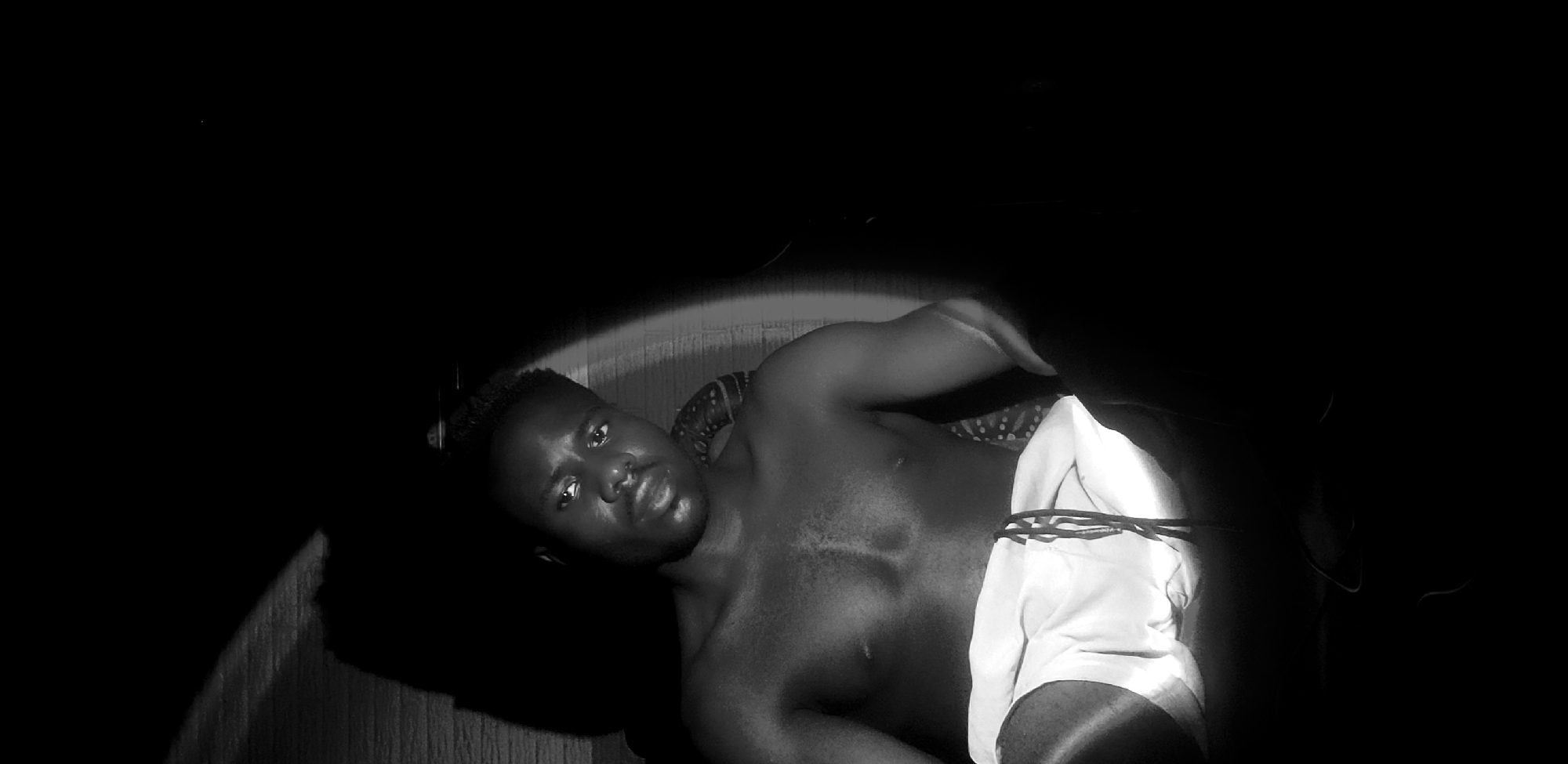 A black and white medium shot captures a shirtless Black man from the chest up, wearing white shorts with a drawstring. He is seated, with his left arm resting on what appears to be a patterned fabric. Dramatic lighting casts a strong shadow from the left, illuminating his face and upper body with a spotlight effect. The background is dark, with a textured wall visible behind him on the left, where the shadow originates. The man's gaze is directed slightly off-camera to his left. The scene appears to be indoors, possibly in Narayi, Nigeria, given the context.
