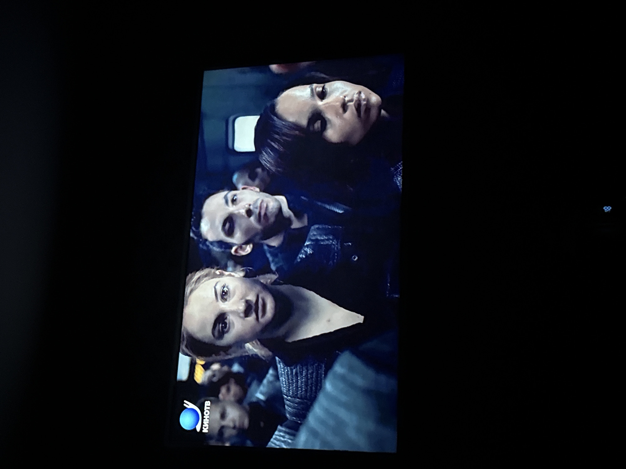 A photograph shows a scene on a television screen, which includes three people in the foreground. The lighting is subdued. The photograph is somewhat rotated counter-clockwise. The people look like they are in a crowded space or vehicle. The logo of a television network "KinoTB" is at the bottom of the screen. It is unlikely the image location is in Mytishchi, Russia.