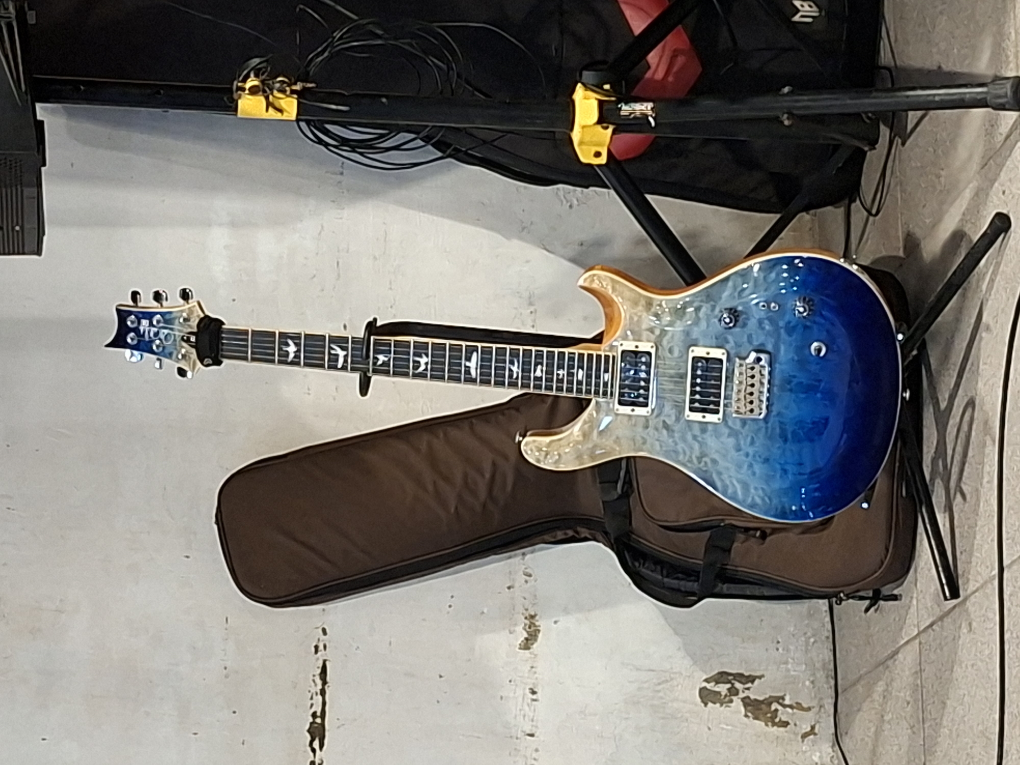 The image shows an electric guitar lying on a brown guitar case, which is placed against a textured wall. The guitar is a blue-to-clear ombre color with a light-colored binding. It is resting on a black guitar stand. Above the guitar is a black stand with yellow clamps and a collection of cables. The location in Lhokseumawe, Indonesia is not discernible from the image.